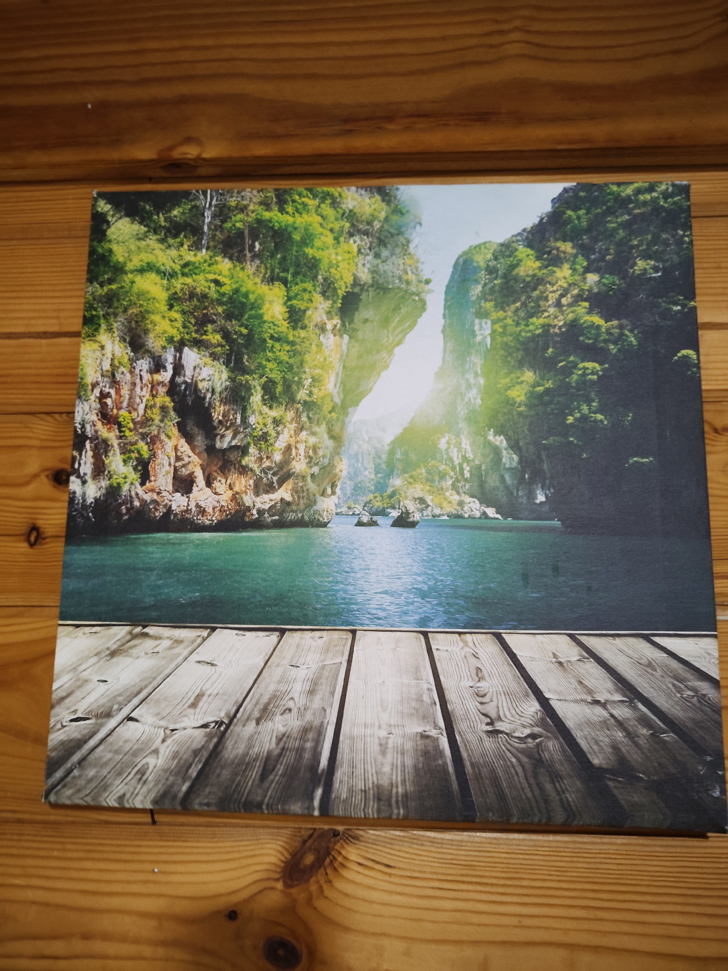 This is a photograph of a canvas print featuring a tropical landscape. The print depicts a body of turquoise water with lush green cliffs rising on either side, creating a narrow passage. Sunlight streams through the opening between the cliffs, casting a glow on the water and vegetation. In the foreground of the print, there is a wooden pier made of planks, giving the impression of standing on a dock overlooking the scene. The canvas print is placed on a surface of horizontal wooden planks, likely a wall or furniture, with visible wood grain and knots. The overall impression is of a serene and picturesque natural setting. There are no people or animals visible in the image. The location in the city of Фестивальное Снт, Russia is not depicted or suggested by the image.