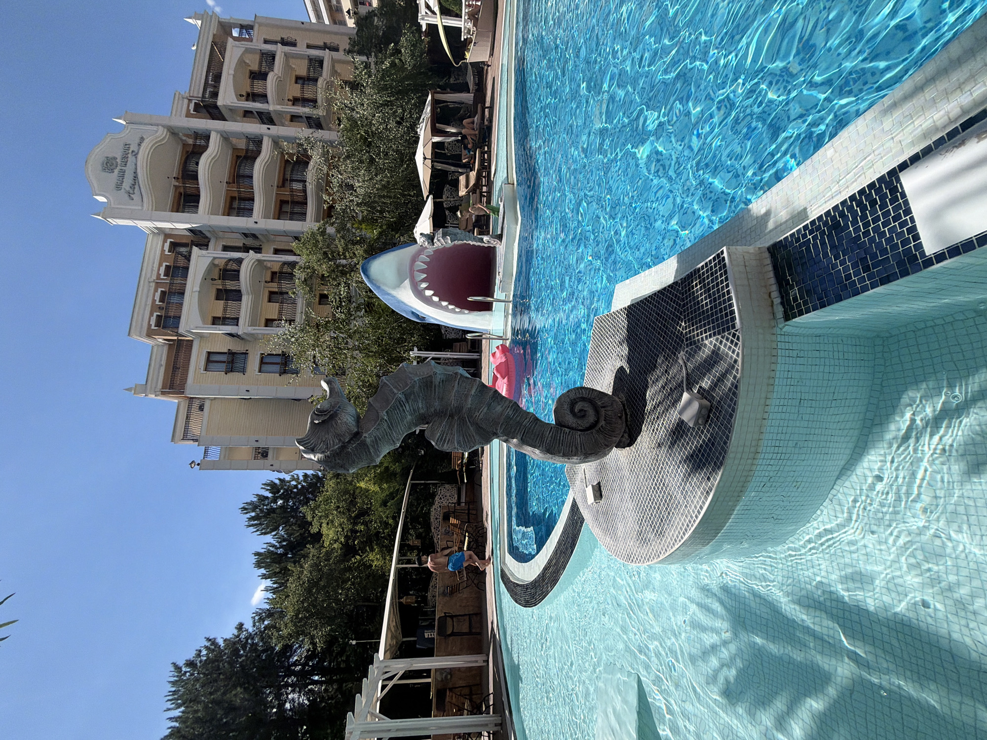 The image depicts a resort in Nesebar, Bulgaria, featuring a large, bright blue swimming pool. In the foreground, a dark, coiled statue of a seahorse stands on a tiled platform in the pool. To the right, a large, cartoonish shark mouth serves as an entrance to a water slide, with its jaws wide open. A pink inflatable ring floats in the pool near the shark. In the background, a multi-story hotel building with balconies and a decorative facade rises above lush green trees. A man in blue shorts is visible near a covered bar area to the left. Several lounge chairs and umbrellas are arranged around the pool, with some people relaxing on them. The sky is clear and blue, indicating a sunny day. The water in the pool is clear, with sunlight creating rippling patterns on the tiled bottom.