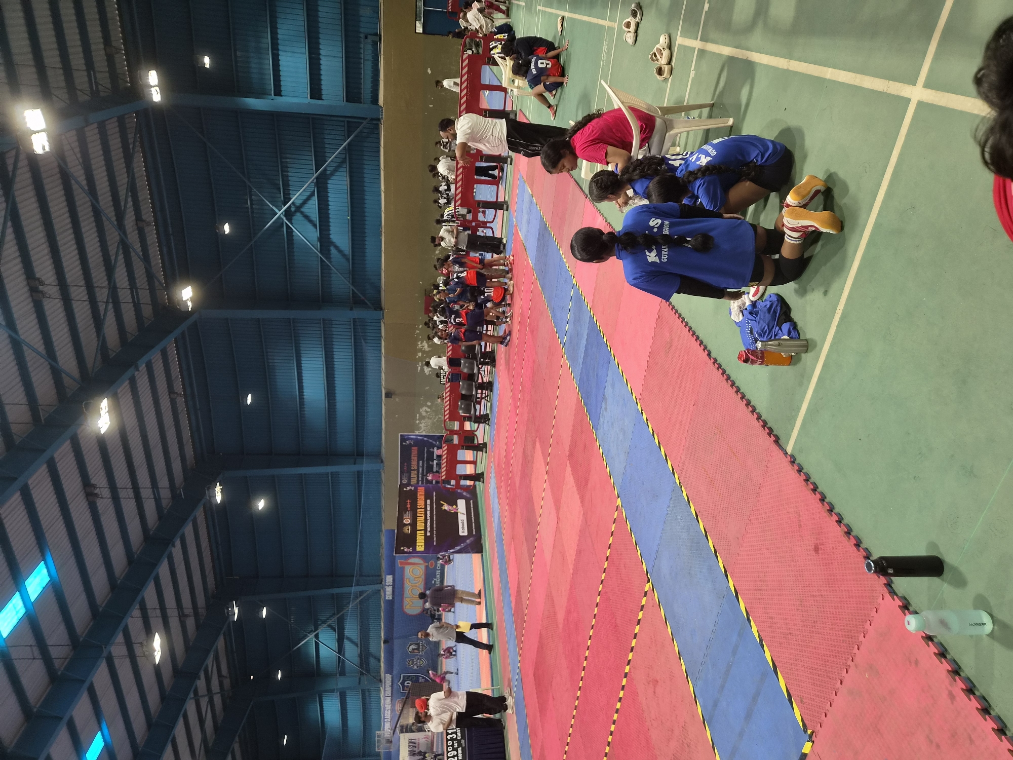 The image depicts an indoor sports arena, likely a gymnasium, in Phagwara, India, filled with participants and spectators for a sports event, possibly a kabaddi match given the branding on a banner. The floor is covered with interlocking foam mats in red and blue, separated by yellow and black striped tape. Several individuals, predominantly young women in athletic attire, are visible on and around the playing area, some actively participating, others resting or observing. Banners displaying event information, including "Kendriya Vidyalaya Sangathan 60th National Sports Meet 2023," are displayed on the back wall. Spectators are seated on chairs along the sidelines, and some individuals are standing, observing the proceedings. The arena is illuminated by overhead lights, and the ceiling structure is visible. The overall atmosphere suggests a competitive sporting event in progress.