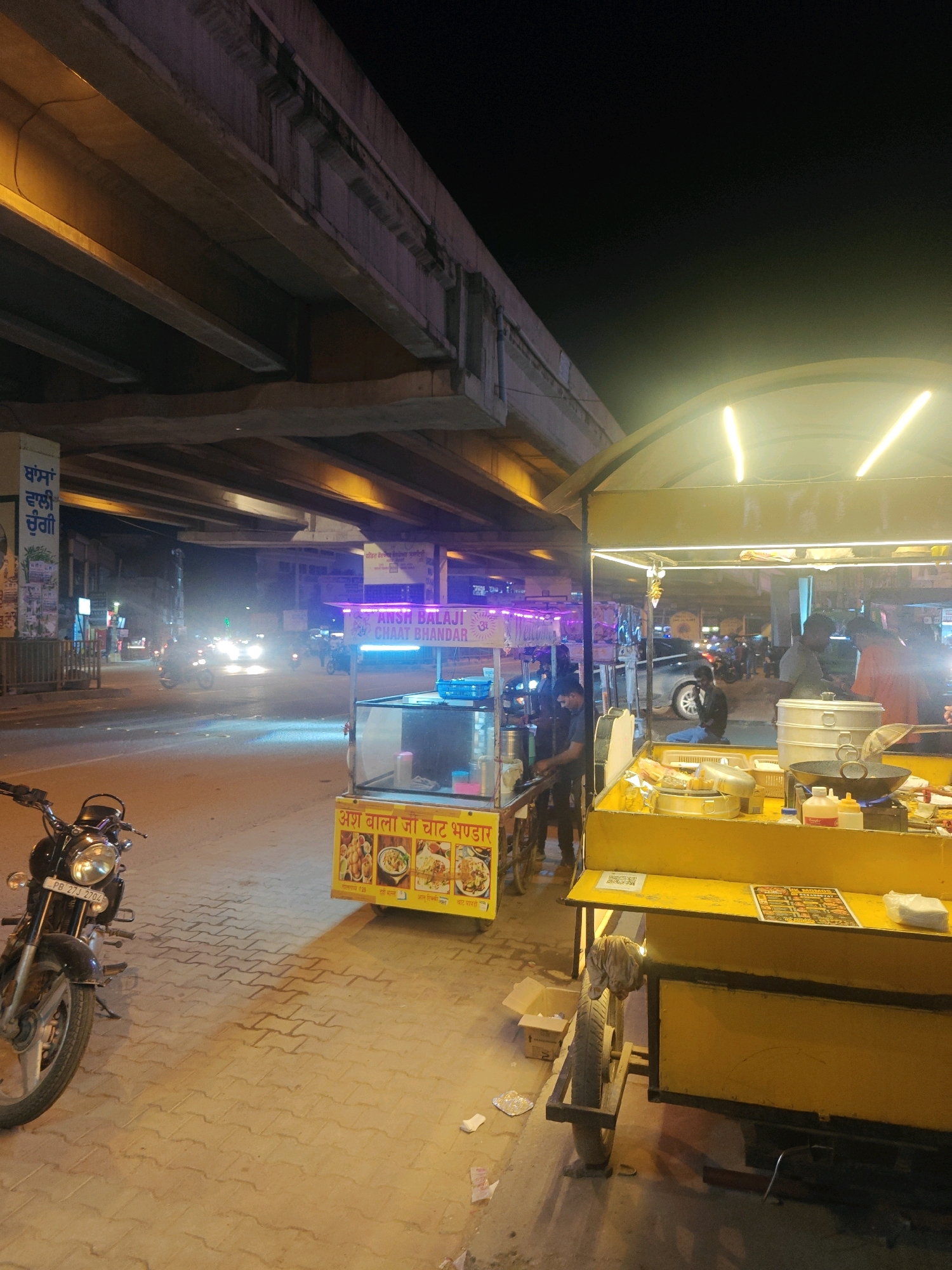 The image depicts a nighttime scene in Kharar, India, featuring a street food stall operating beneath a large overpass. The stall, illuminated by bright yellow lights, displays various food items, likely chaat and other snacks, on its counter. A wok is visible on a burner, suggesting food preparation. Several individuals are present around the stall, with some appearing to be customers and others working. A motorcycle is parked to the left of the frame, and a second, smaller food cart is visible to the left of the main stall. The street is paved with interlocking bricks. The overpass looms overhead, with illuminated sections indicating traffic. The overall atmosphere is that of a busy urban night market.