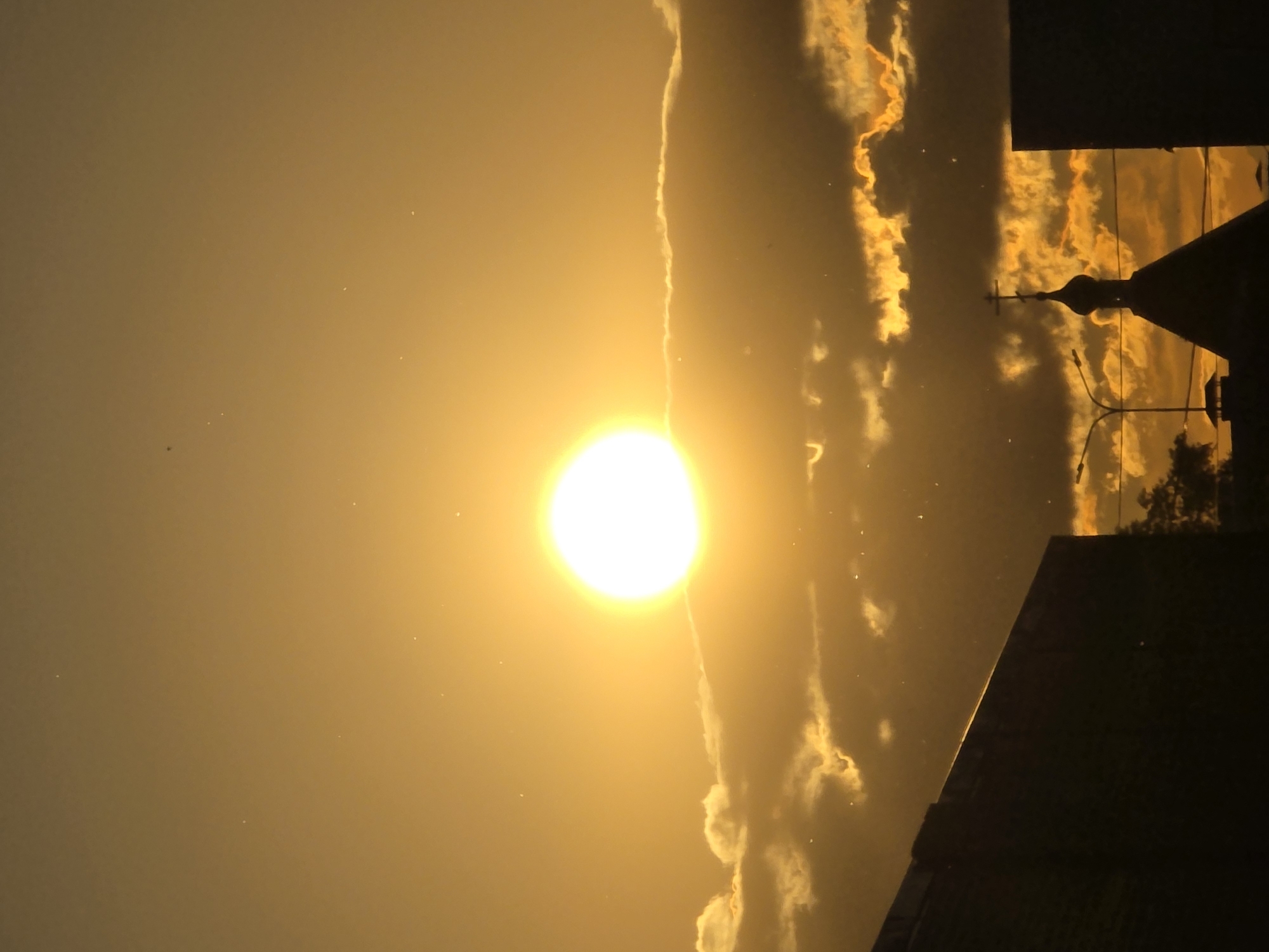 This media file captures a vivid sunrise or sunset scene over what appears to be an urban or semi-urban landscape in Chaikovsky, Russia. The dominant feature is the brilliant, overexposed white disc of the sun, positioned centrally on the left side of the frame. The sky is bathed in intense golden and orange tones, transitioning from a lighter hue near the sun to a deeper golden color further away.
Scattered clouds are visible, particularly on the right side of the image, illuminated from behind by the sun's powerful light, creating bright, glowing formations against the darker sky. A prominent silhouette of what appears to be an Orthodox church dome topped with a cross is visible on the right side of the image, strongly hinting at the Russian location. Alongside this, other dark silhouettes of buildings and possibly utility poles or streetlights are discernible against the glowing sky.
Numerous small, bright specks are scattered across the sky, particularly in the areas not directly obscured by the sun's glare. These are likely dust motes either in the atmosphere or on the camera lens/sensor. The low angle of the sun and the dramatic golden lighting firmly establish the time of day as either early morning or late evening. There is no visible text in the image. The scene evokes a sense of natural beauty and the serene grandeur of a celestial event.