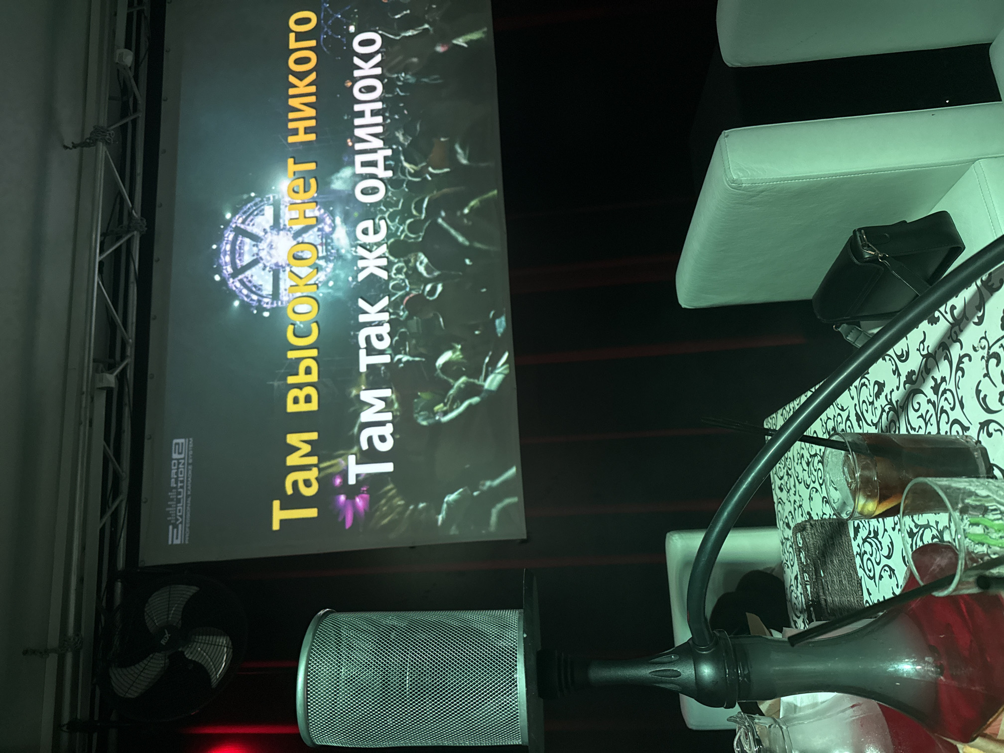The image shows a karaoke setup inside a bar or club, likely in Chaikovsky, Russia, with lyrics displayed on a screen reading "Там высоко нет никого. Там так же одиноко." (There is no one up there. It is just as lonely there.) A microphone is positioned in the foreground. Several drinks in glasses are on a table with a black and white pattern. A white cushioned seating area is in the background. A black fan and the Evolution Pro 2 Karaoke System logo are visible on the screen.