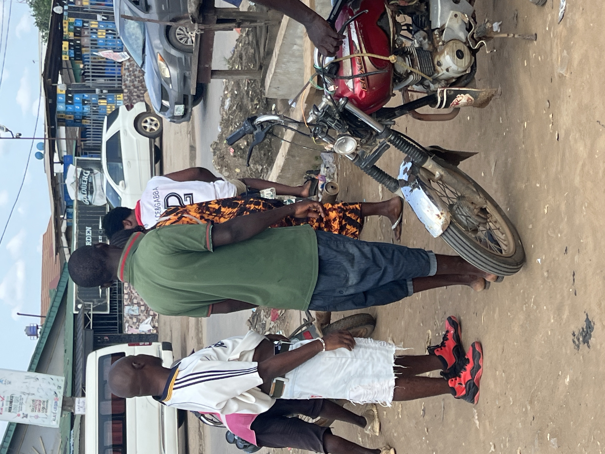 The image captures a street scene in Ijegun, Nigeria. A red motorcycle is parked on the side of the road. Several people are standing nearby. One person is wearing a green shirt and dark shorts. Another person is wearing a white and black shirt, white shorts, and red and black sneakers. In the background, there are buildings, cars, and stacks of blue crates.