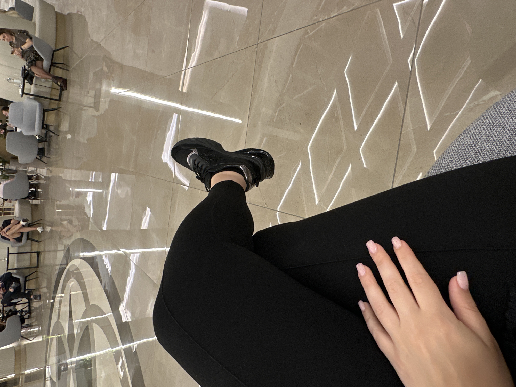 The image, taken in Kazanlak, Bulgaria, is a first-person, low-angle shot looking down at the subject's legs, feet, and left hand. The person is seated, wearing black form-fitting leggings and chunky black sneakers with silver accents. Their left hand, resting on their lap, features light pink manicured nails.
The setting is a sleek, modern indoor public space, likely a lounge, cafe, or hotel lobby, characterized by highly polished light-colored tiled floors that create numerous reflections. The floor design is intricate, featuring geometric patterns composed of bright, narrow lines—some forming diamond shapes, others large concentric arcs. These lines appear to be integrated lighting or highly reflective elements, casting a luminous glow across the surface.
Reflected in the glossy floor are several other individuals seated at small tables with grey chairs. One woman, wearing a patterned dress, is clearly visible in the reflection, appearing to be engaged with something in her hands on a table with bottles and glasses. Other blurry reflections of people and furniture are also visible, contributing to the sense of a busy yet sophisticated environment. The predominant activity for the visible subjects is sitting and waiting or socializing. The lighting appears to be artificial, typical of an indoor setting, with no indication of the time of day or outdoor weather. There is no visible text.