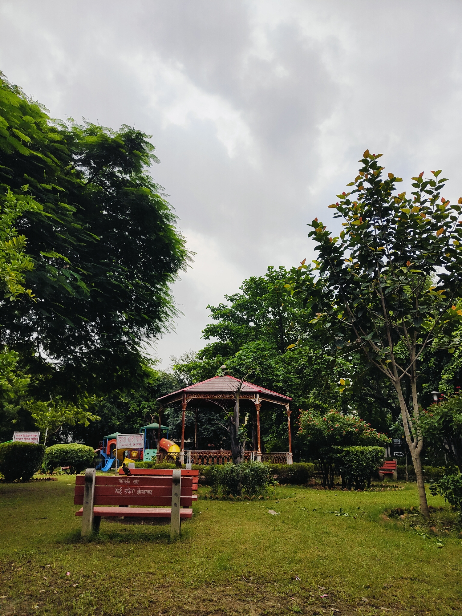 The image depicts a park scene in Gurgaon, India, with lush green grass, mature trees, and a playground. In the foreground, a red bench with concrete legs is visible, with text inscribed on its backrest in Hindi. Behind the bench, a children's playground with slides and other colorful equipment is situated. A decorative gazebo with intricate metalwork stands further back, surrounded by trees and bushes. The sky above is overcast with grey clouds. The scene appears peaceful and well-maintained, suggesting a recreational area within a city park.