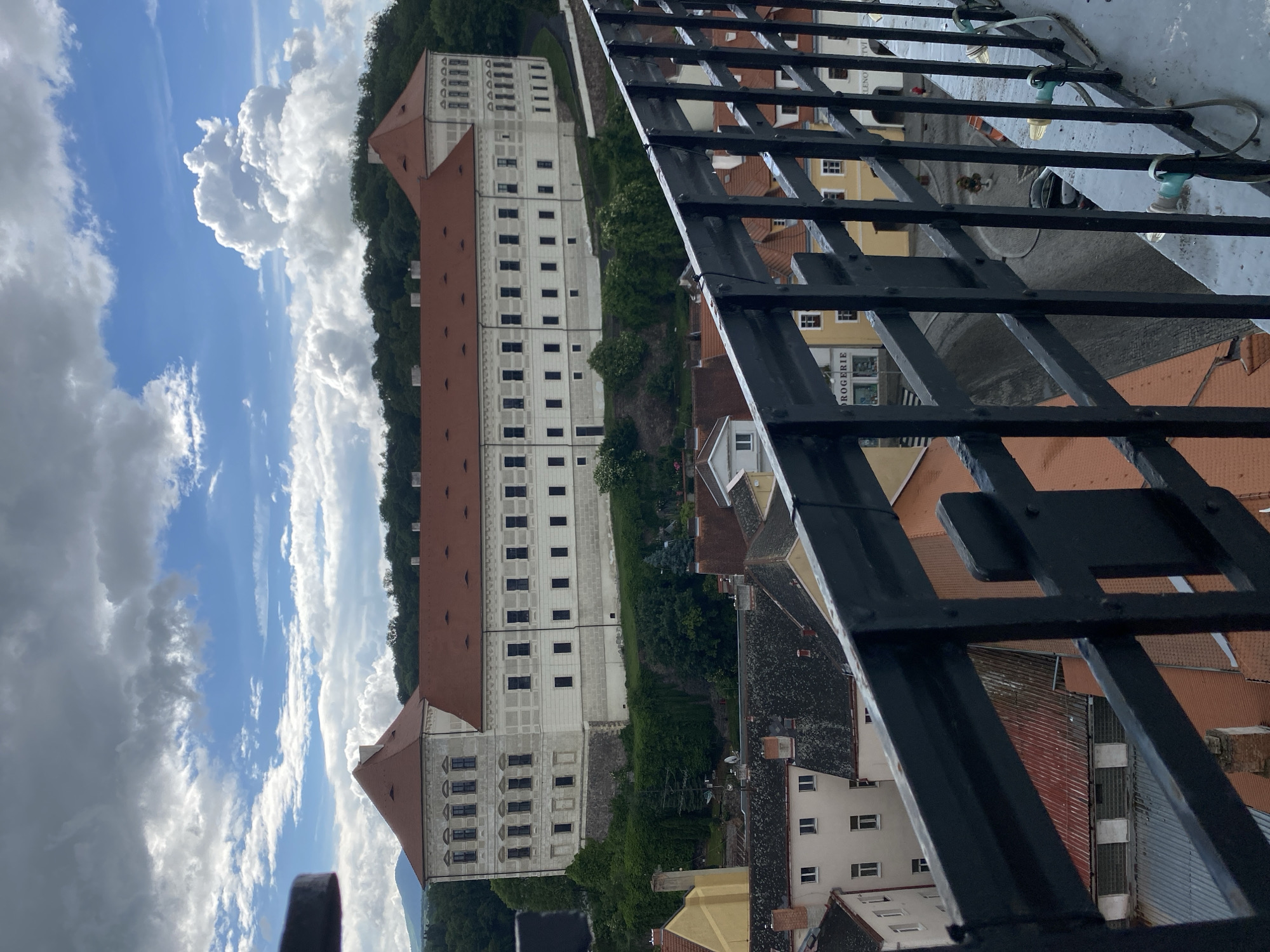 The image shows a view of the Bílina Castle in Bílina, Czechia, from a higher vantage point, possibly a balcony or observation deck, looking over a town with red-tiled roofs. The castle is a large, imposing structure with a white facade and red-tiled roofs, situated on a hill covered in green trees. The sky is partly cloudy, with dramatic white cumulus clouds against a blue background. In the foreground, a black metal railing with lights attached obstructs the view of the town below. The town itself consists of several buildings with orange and red roofs, and some yellow and white facades. A street is visible with a few people and a parked car. The overall scene depicts a scenic view of a historic European town and its prominent castle.