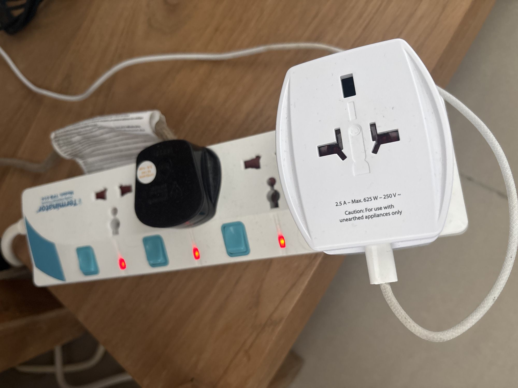 The image shows a close-up, high-angle shot of a white power strip with multiple outlets and individual switches. The power strip is placed on a wooden surface. A black electrical adapter is plugged into one of the outlets, and another white adapter is plugged into an adjacent outlet, with a white cable extending from it. Red indicator lights are visible on the power strip, suggesting it is active. The environment appears to be indoors, possibly a home or office. There are no people visible in the image. The overall scene is clean and well-lit, with the focus on the electrical devices. No elements in the image suggest a specific location within Dubai, United Arab Emirates, or any other specific geographical region.