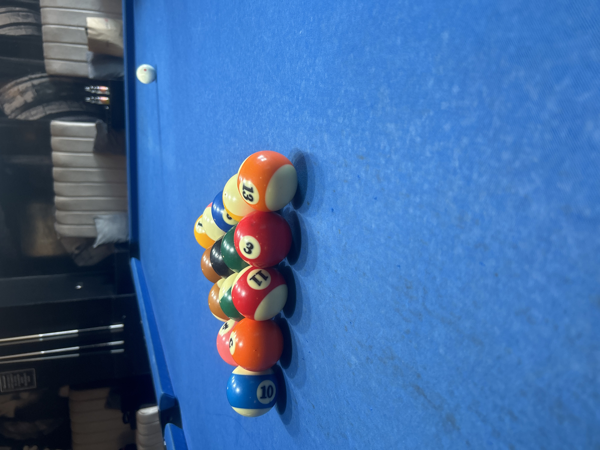 An eye-level, slightly angled indoor shot captures a portion of a billiards table in Pleven, Bulgaria. The table is covered with a textured, light blue felt surface, exhibiting some minor wear. A white cue ball is visible near the top-left edge of the frame.
The primary focus is on a collection of billiard balls clustered in a somewhat diagonal arrangement on the blue felt, extending from the middle-left towards the bottom-right. The visible balls include a mix of solid and striped types. Clearly identifiable solid balls are: a red ball with the number '3', a red and white ball with '11', an orange and white ball with '13', a yellow and white ball with '15', and a blue and white ball with '10'. Among the striped balls, a yellow and white ball with '9' and a green and white ball with '14' are discernible. Partially visible balls include a dark green solid ball and a brown solid ball.
The background on the left side of the image is out of focus, depicting indistinct elements. These blurry shapes suggest stacked furniture or storage units in light tones, along with a dark, possibly wooden, cabinet or wall paneling. Hint images reveal cue sticks stored in a rack against this dark background. The lighting in the scene appears to be artificial, typical of an indoor setting like a game room or bar. No individuals are present in the image. The arrangement of the balls suggests a game of billiards is either in progress or set to begin.