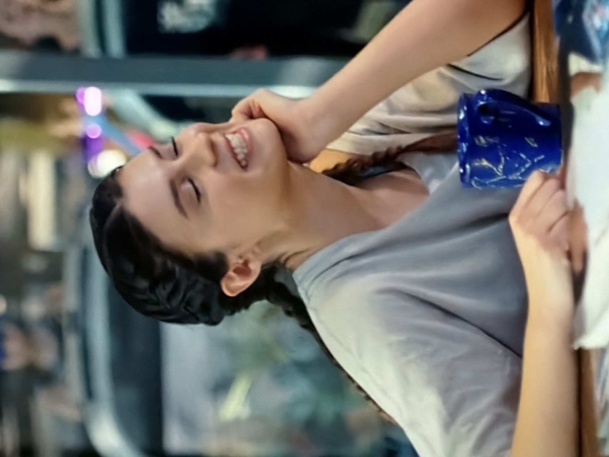 A young woman with dark hair styled in braids is sitting at a table, smiling with her eyes closed. She is wearing a light-colored t-shirt. Her right hand is resting on her chin. In front of her is a blue mug with a textured design. The background is blurred, showing glimpses of people and what appears to be a café or restaurant setting, possibly in Kazan, Russia, indicated by the lighting and general ambiance.