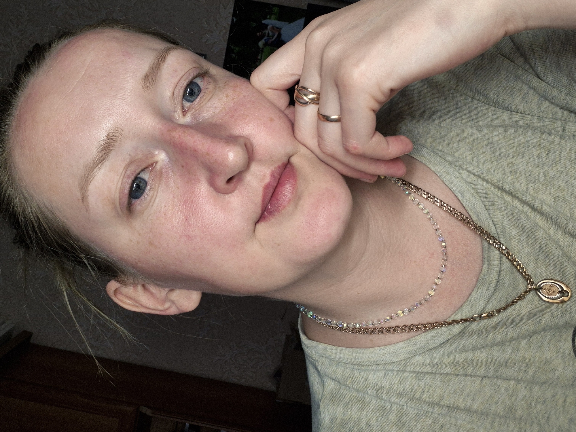 A close-up portrait of a fair-skinned woman with freckles and blue eyes is captured indoors. She is wearing a light green t-shirt and two delicate necklaces, one with iridescent beads and the other with a golden chain and pendant. Her left hand, adorned with two rings, rests against her face. The background appears to be a dimly lit room, and a framed picture hangs on the wall behind her. There is no information in the image to verify if the location is Chaikovsky, Russia.