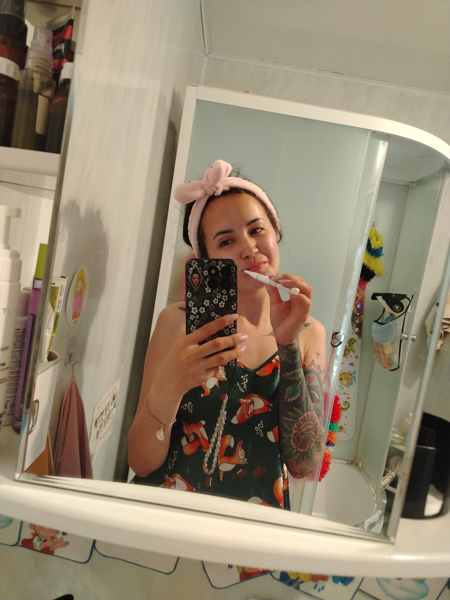 A woman stands in a bathroom, reflected in a mirror. She is wearing a pink headband and a patterned top with foxes. She holds a black smartphone in her left hand and a white object resembling a thermometer or pen in her right hand, near her lips. Her left arm is covered in tattoos. The bathroom features a cabinet with toiletries, a towel, and a shower stall with accessories hanging inside. The mirror frame has a patterned border. No information exists in the image to verify the location as Noviy, Russia.