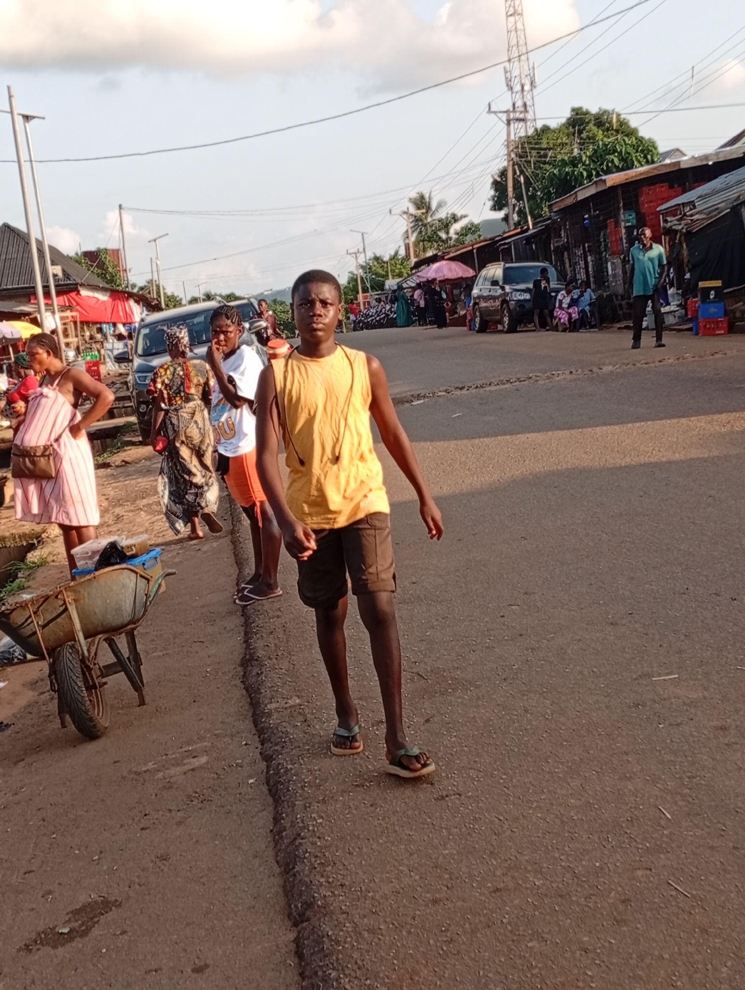 In Nsugpe, Nigeria, a bustling roadside market is captured. A young Black male, wearing a yellow sleeveless shirt and brown shorts, walks toward the camera, his right hand slightly raised. To his left, a Black female, dressed in a pink and white striped dress and carrying a brown purse, stands with her hands on her hips. Behind her, another Black female in a white t-shirt and orange shorts faces away, while a third Black female in a patterned dress is also visible. A two-wheeled wheelbarrow with a metal basin is positioned to the left of the frame, near the edge of the road. In the background, various market stalls with corrugated metal roofs line the street, with vehicles, including a dark SUV, parked nearby. Overhead, power lines crisscross the sky, and a cell tower is visible in the distance. The scene suggests a typical day at a Nigerian market, with people engaged in commerce and daily life under a bright, partly cloudy sky.