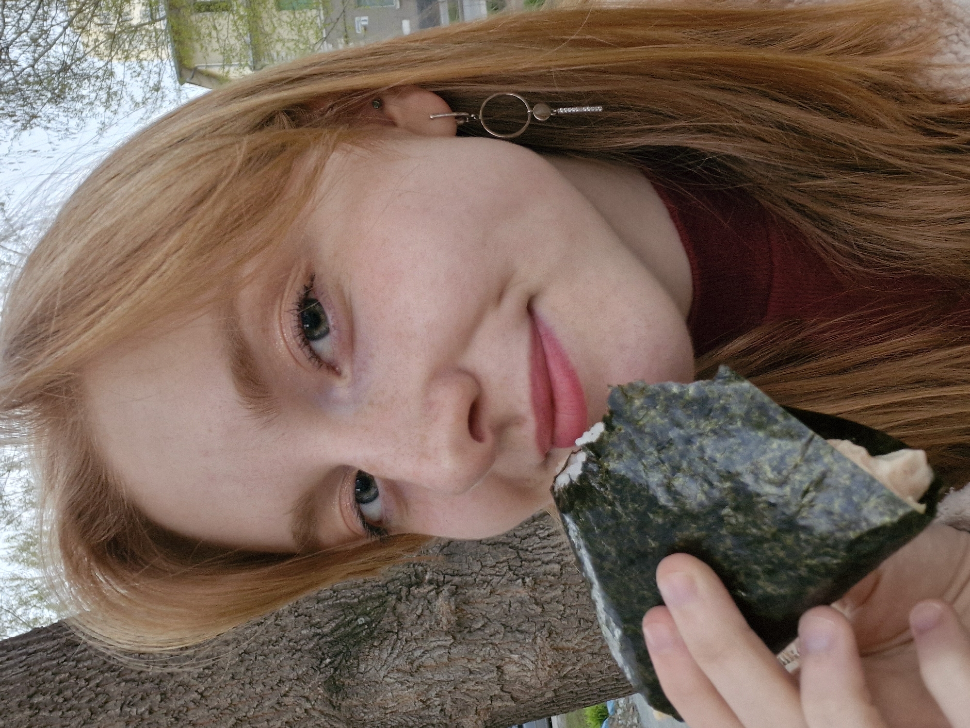 A casual, close-up portrait of a young individual is captured at an angle, tilting the scene counter-clockwise. The subject, likely female, has fair skin with visible freckles across their nose and cheeks, and striking light-colored eyes. Their long, reddish-blonde hair frames their face and extends into the background, partially obscuring a dark red top or sweater. They wear a distinctive silver earring on their visible ear, featuring an open circle, a small dot, and a bar adorned with small, sparkling elements.
The individual is engaged in eating, holding a piece of food resembling an onigiri or sushi roll, wrapped in dark green nori seaweed. The food appears partially bitten or opened, revealing a light-colored filling inside. Their lips are slightly parted near the food, and they look towards the camera with a gentle smile or relaxed expression.
The background suggests an outdoor urban or suburban setting, possibly in Yekaterinburg, Russia. A textured tree trunk occupies the lower portion of the frame. Above it, blurred elements of distant buildings and sparse green foliage are visible under a bright, overcast or light sky, indicating it is daytime. The overall image conveys a relaxed, everyday moment. There is no visible text.