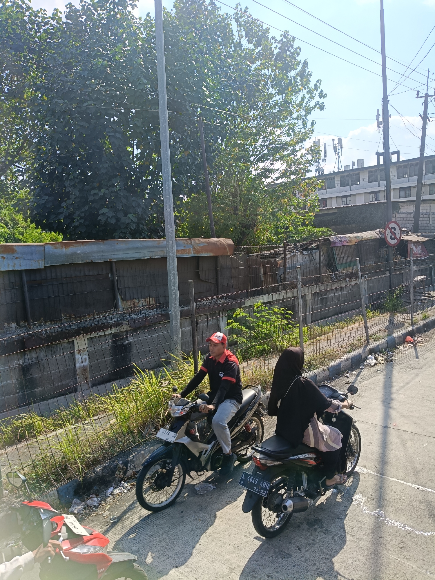 Two individuals are riding motorcycles on a paved road adjacent to a drainage ditch in what appears to be an urban or semi-urban area. The person in the foreground on the left is wearing a red cap and a black and red shirt. The person on the motorcycle behind them is wearing a black hijab and a black outfit. Behind the motorcycles, there is a metal fence, followed by a concrete wall and then a row of small, makeshift structures with corrugated metal roofs. Lush green vegetation is visible growing along the ditch and behind the structures. In the background, there are buildings, power lines, and cell towers, suggesting a developed area. The sky is clear and blue with some scattered clouds. The scene is lit by bright sunlight, casting shadows on the road. The license plate on the rear motorcycle reads "L 4649 ABH" and "09-23". The location is identified as Kedungrejo, Indonesia.