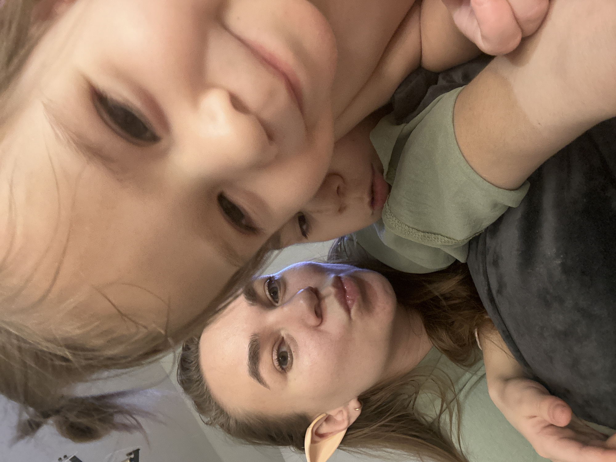 Three individuals are visible in a close-up selfie. At the top of the frame is a young child with light hair and fair skin. Partially obscured beneath them is another young child. At the bottom of the frame is an adult female with light brown hair, fair skin, and light eyes. The adult is wearing a prosthetic pointed ear. The setting appears to be indoors, with a light-colored wall or surface visible in the background. The location is unknown; therefore, the specific city cannot be determined. The individuals appear to be posing for the selfie.