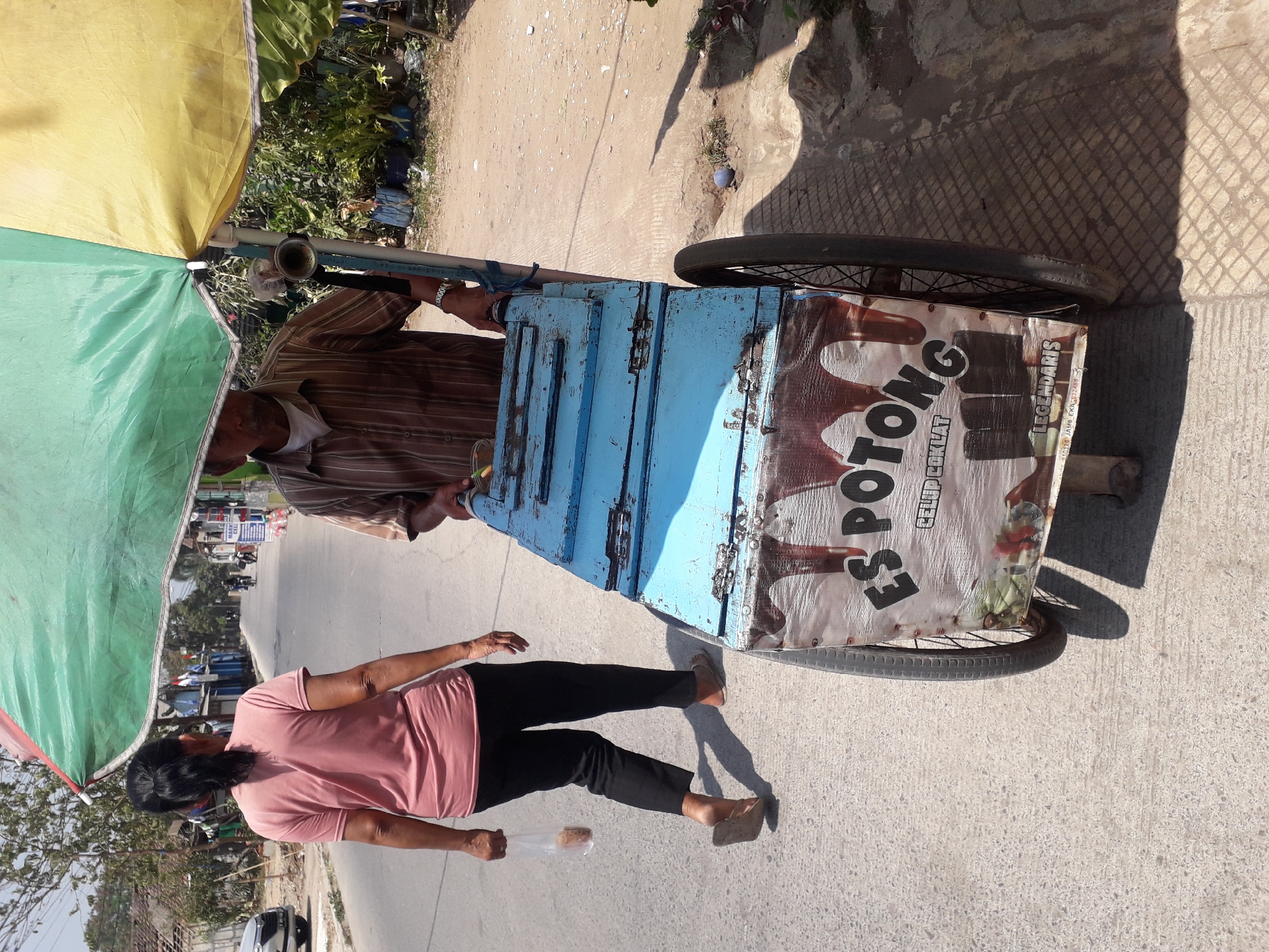 An overcast day in Cankuang, Indonesia, features a street scene with an ice cream cart and two individuals. A woman, wearing a pink t-shirt and black trousers, walks away from the camera with a plastic bag in her left hand. Beside her, an ice cream vendor, dressed in a striped short-sleeved shirt, stands near his cart. The cart, painted a faded blue, has a large umbrella providing shade. The side of the cart displays "ES POTONG" and "LEGENDARIS" in large lettering, along with an image of chocolate. The street is paved, with a dirt and gravel shoulder to the right, lined with foliage. The background shows buildings and trees, suggesting a residential or commercial area.