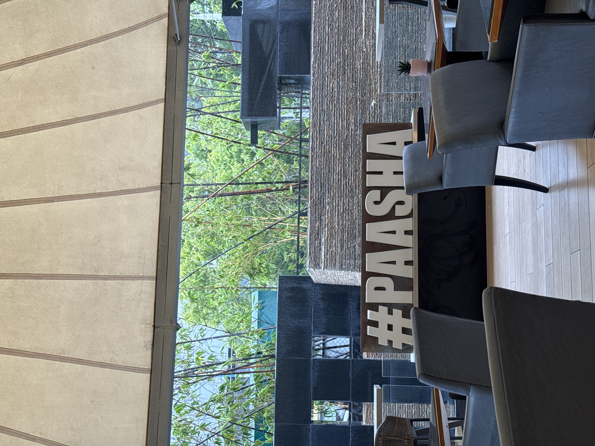 The image captures an outdoor seating area, possibly a cafe or restaurant, with a large "#PAASHA" sign prominently displayed. The sign is made of large, white, block letters on a brown background with a wooden frame. In the foreground, several dark gray upholstered chairs are visible, arranged around tables. A small potted plant sits on one of the tables in the background. Behind the seating area, there's a textured stone wall and a contemporary architectural feature made of dark rectangular blocks. Beyond this, lush green bamboo stalks and foliage are visible, suggesting an outdoor setting with ample greenery. The overall impression is one of a modern, well-designed outdoor space, possibly in a hotel or resort. The location is identified as Koregaon Park, Pune, India, which is known for its upscale restaurants and establishments.