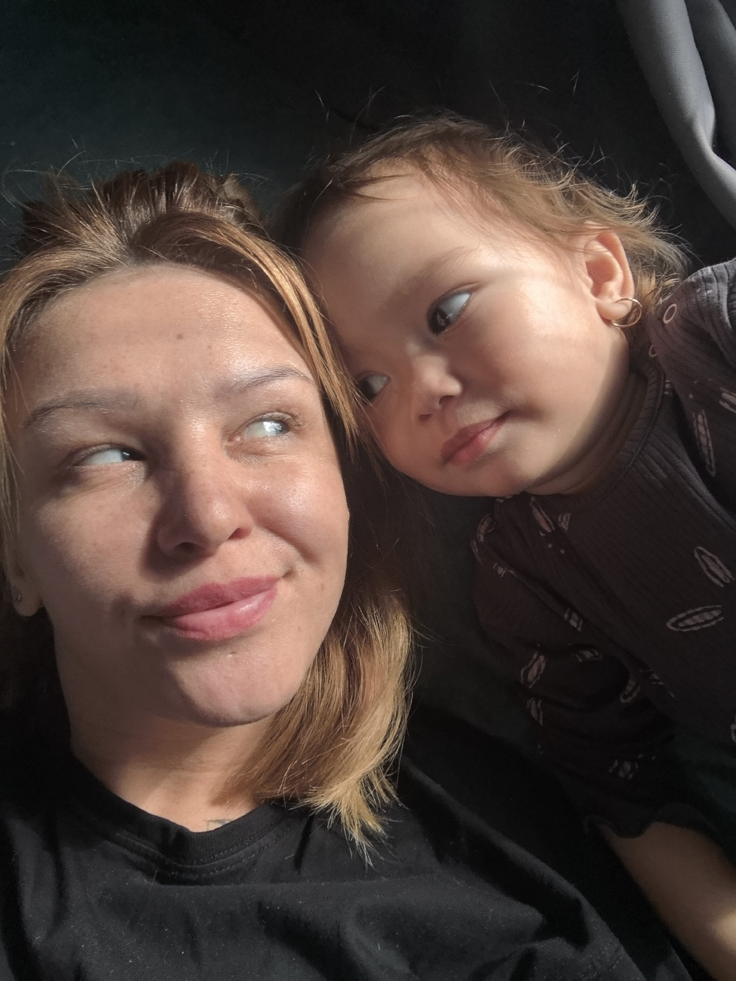 A selfie shows a woman and a young child. The woman has light brown hair and is wearing a black t-shirt. She is looking to her left with a slight smile. The child, who appears to be a toddler, has light brown hair, a gold earring, and is wearing a dark-colored, long-sleeved shirt with a pattern of light-colored oval shapes. The child is looking towards the viewer. The background is dark, suggesting they are indoors. There is no indication of the location being Noviy, Russia.