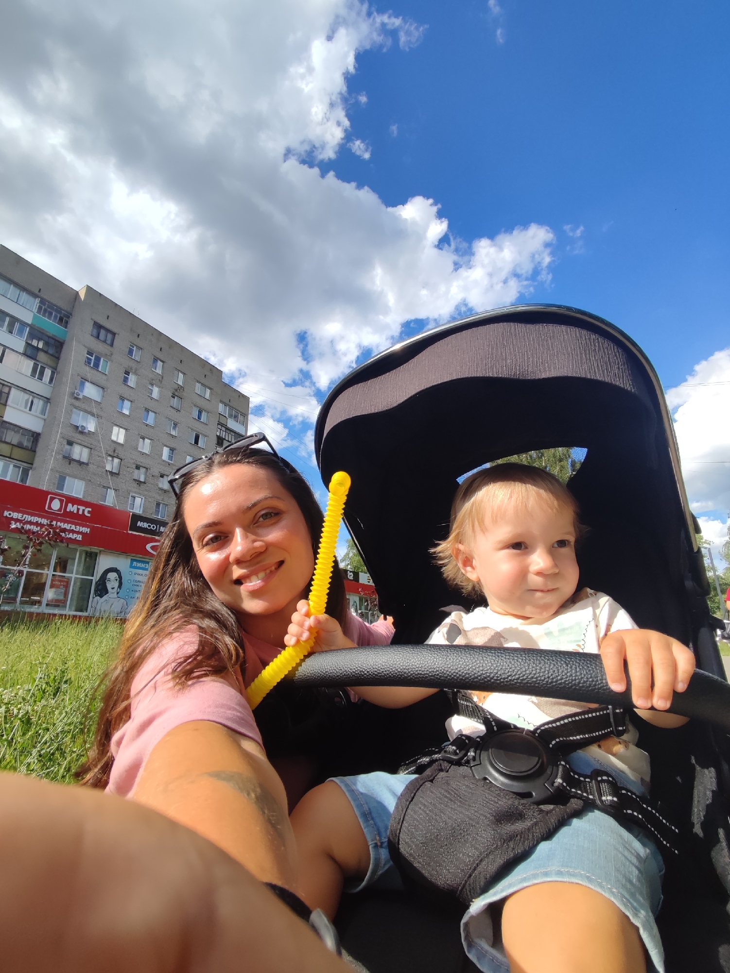A woman and a young child are in a stroller on a sunny day. The woman is smiling at the camera, holding a yellow, flexible toy. The child, in the stroller, is looking to the side. Behind them, a multi-story apartment building and a red building with signs for "MTC" and "МЯСОМ" are visible. The scene is set outdoors, with green grass and a bright blue sky with white clouds. The time of day appears to be daytime. Visible text includes "MTC", "МЯСОМ", and other Cyrillic characters that appear to be store names or advertisements. The overall mood is cheerful and relaxed. The location is Chaikovsky, Russia.