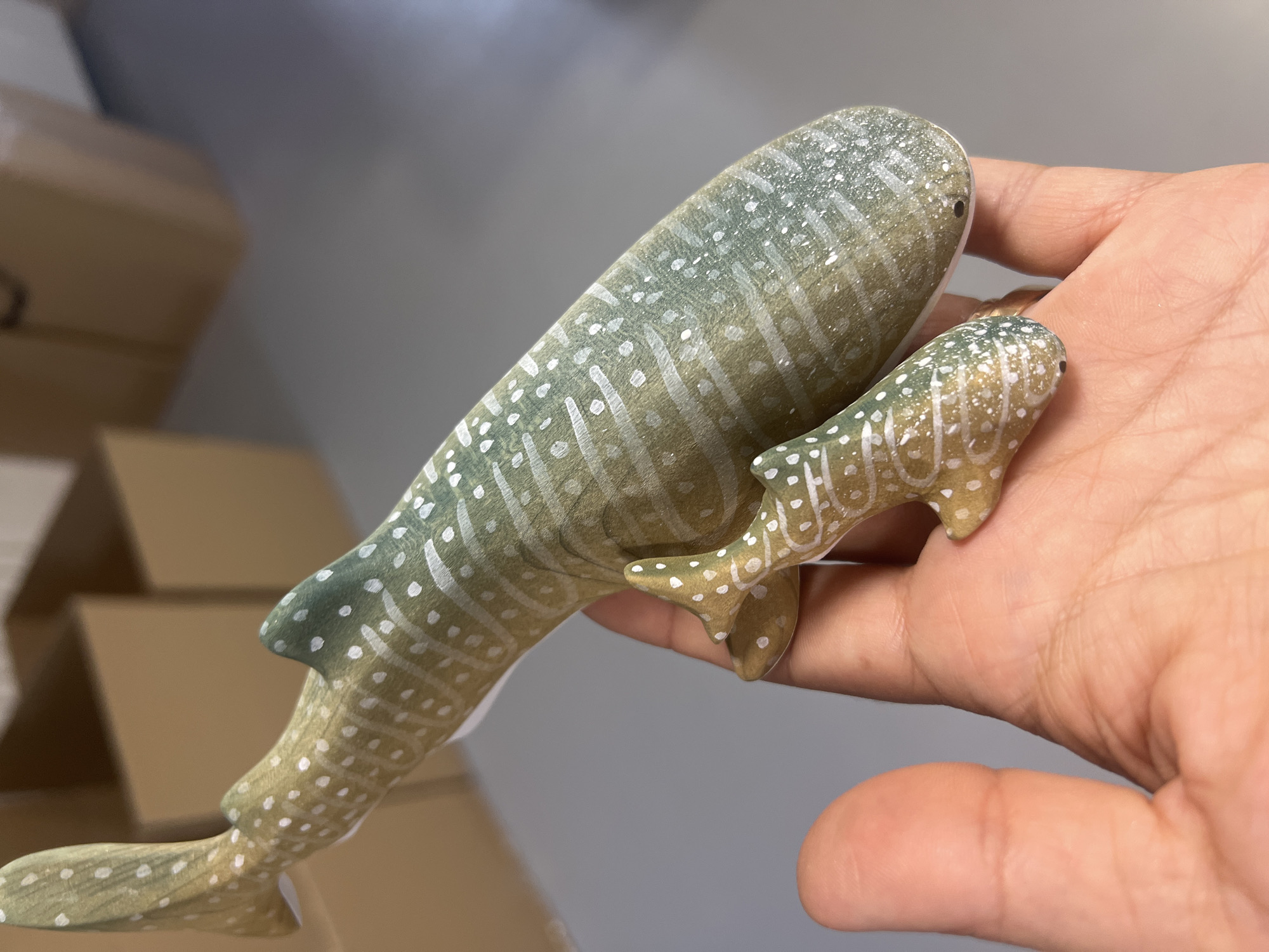 Two decorative wooden figurines of whale sharks, one larger than the other, are being held in a person's left hand. The figurines are painted with a greenish-brown base color and decorated with white spots and wavy white lines. The larger figurine is positioned horizontally, with its tail extending towards the top left of the frame. The smaller figurine is placed in front of the larger one, angled slightly upwards. The background consists of blurred cardboard boxes and a gray floor. The lighting is soft and diffused, suggesting an indoor setting.
