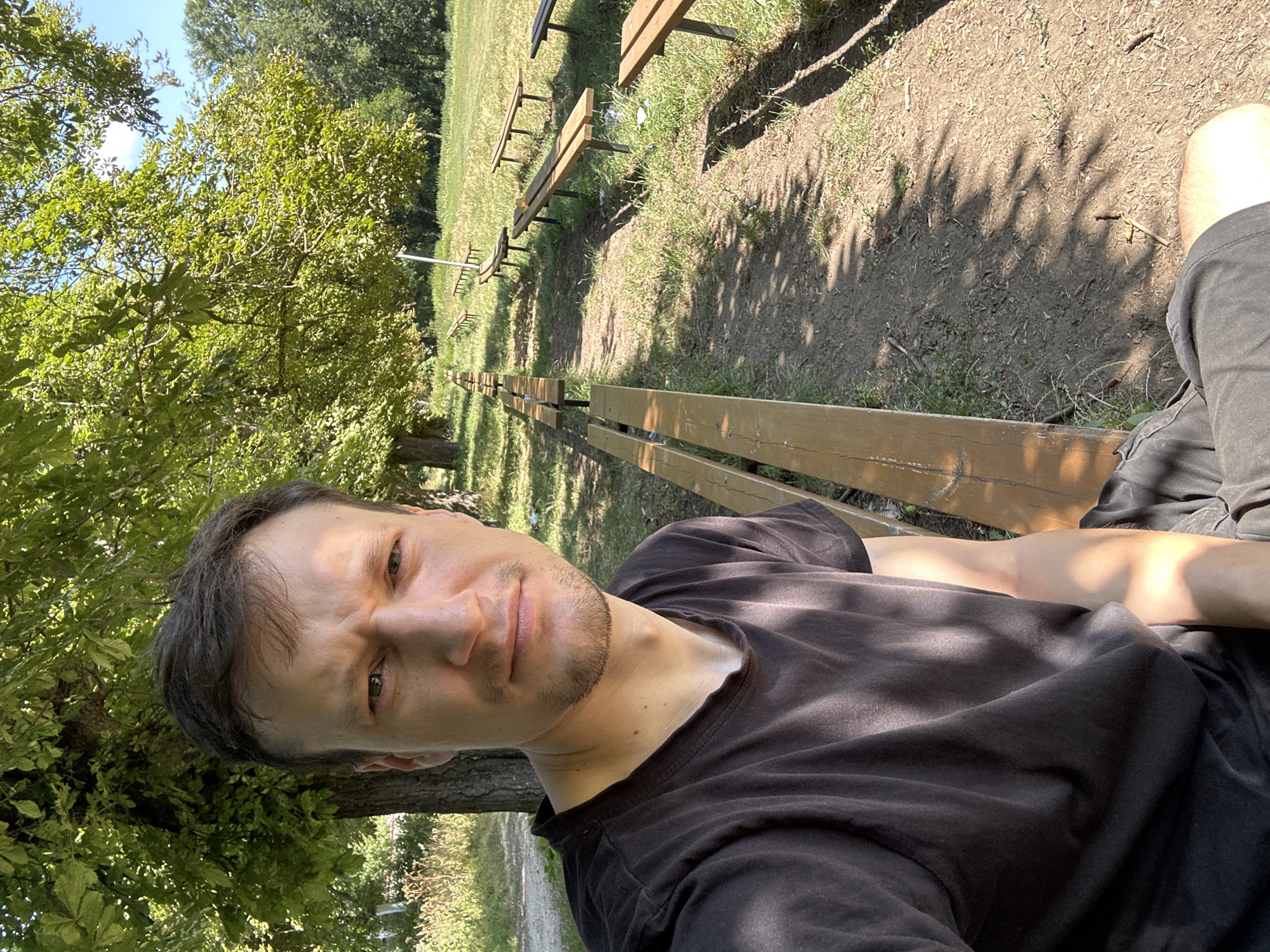 A man is sitting on a wooden bench in an outdoor amphitheater setting. The amphitheater is located in Makiv, Ukraine, and is characterized by rows of wooden benches facing a grassy area. The man is positioned in the foreground, looking directly at the camera with a neutral expression. He is wearing a black t-shirt and dark shorts. Sunlight filters through the leaves of a large tree, casting dappled shadows on the man and the ground. The background shows more rows of benches receding into the distance, surrounded by grass and trees, under a clear blue sky.