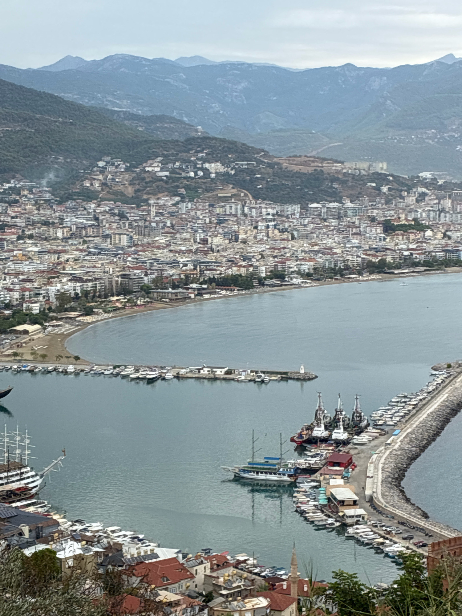 The image captures a panoramic view of the Alanya Marina in Alanya, Turkey, with the city sprawling towards the mountainous backdrop. In the foreground, a cluster of buildings with red-tiled roofs is visible, nestled amongst greenery. The marina itself is a prominent feature, filled with a diverse collection of vessels, ranging from large sailing ships and yachts to smaller fishing boats and speedboats. Several of these boats are moored along a curved, stone-constructed breakwater that extends into the harbor.
The water of the harbor is a muted, bluish-gray, reflecting the overcast sky. Across the harbor, the city of Alanya stretches along the coastline. Numerous buildings, primarily multi-story residential and commercial structures with white and light-colored facades, are densely packed together. The city extends up the slopes of the hills, where some buildings are more scattered.
Beyond the city, a range of imposing, hazy blue mountains forms the horizon. The overall atmosphere is serene, with a sense of a bustling port city integrated into a natural landscape. No people are distinctly visible in the image, contributing to the still and tranquil feel of the scene. The lighting suggests it is daytime, possibly late afternoon or an overcast day, as there are no harsh shadows.
