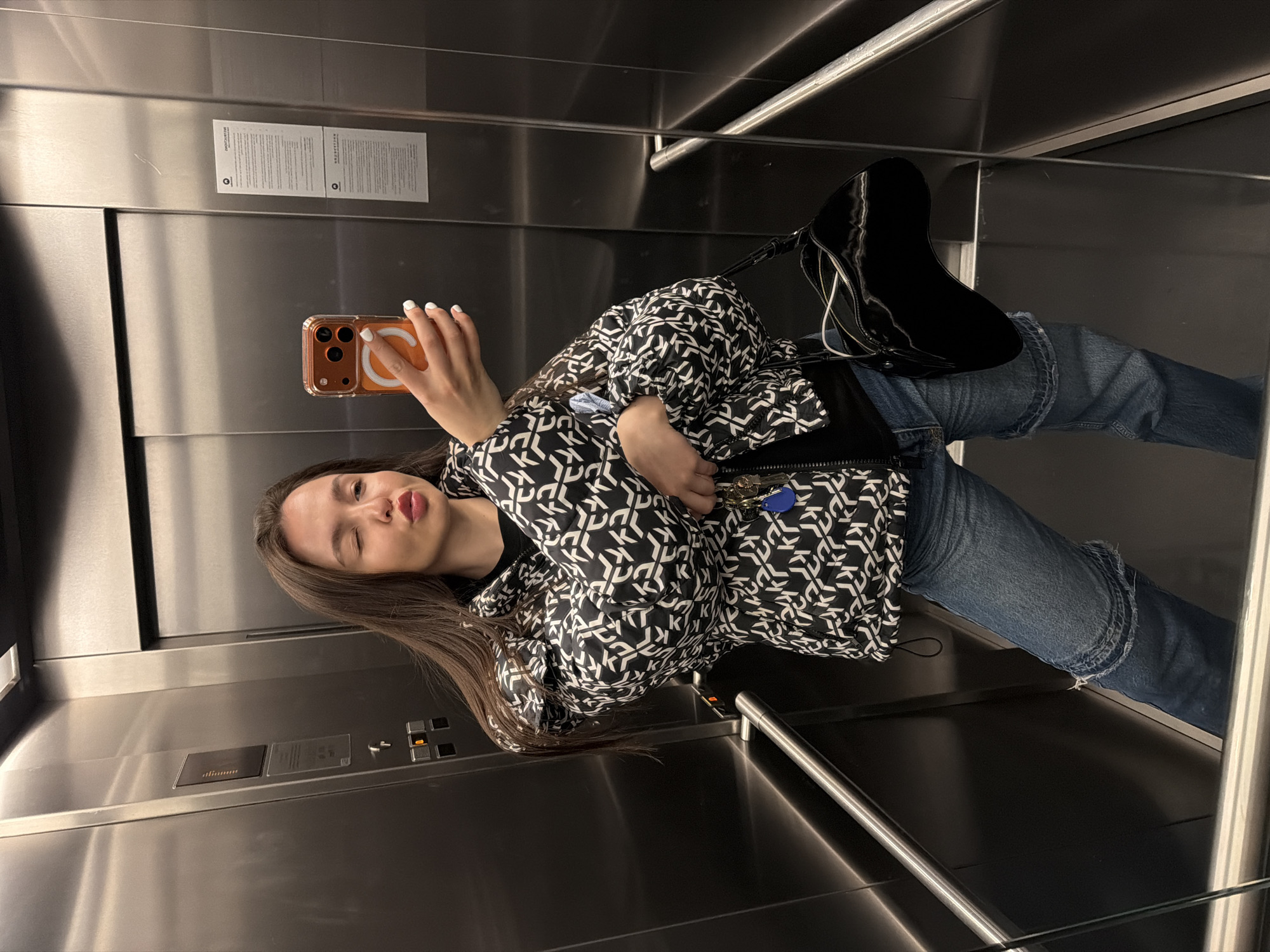 A young woman is taking a selfie in an elevator. She is wearing jeans, a black and white patterned jacket, and black high-heeled boots. She is holding a phone in her right hand with white nails. The elevator has stainless steel walls and handrails. There are elevator buttons and informational signs visible. The image doesn't provide location context for Sofia, Bulgaria.