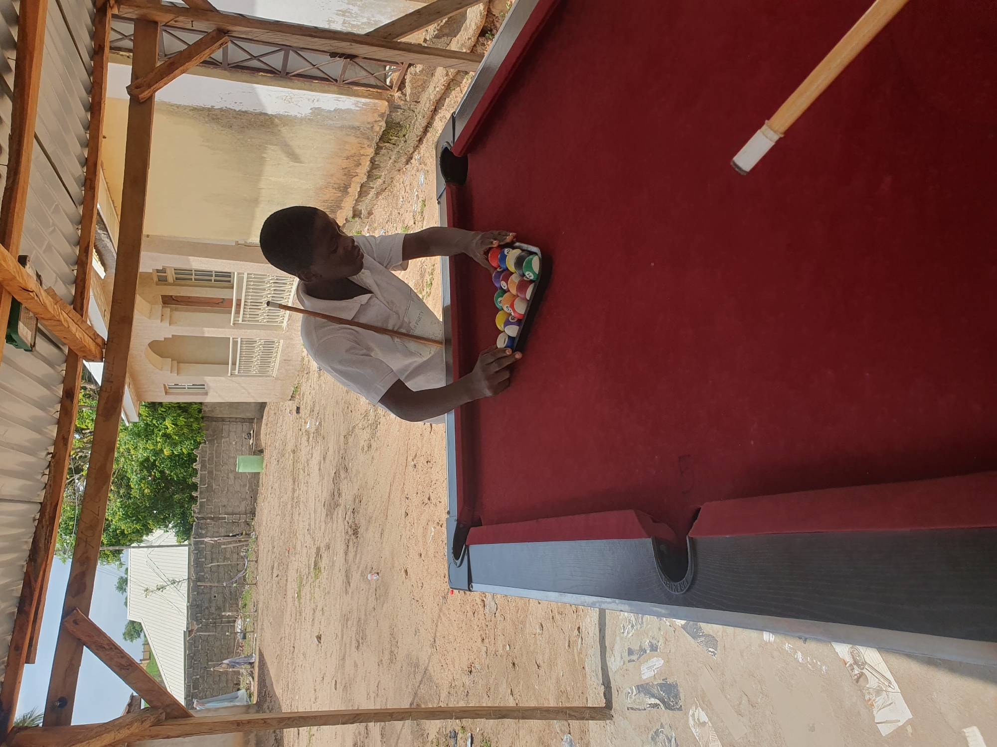 A young Black male wearing a white shirt is shown setting up billiard balls on a red felt billiard table. The balls are arranged in a triangular rack. He is standing outdoors, under a corrugated metal roof supported by wooden beams. In the background, there is a sandy yard with a brick wall, a light-colored building with arched windows, and some trees. The scene appears to be in a somewhat unkempt, possibly residential area. The lighting suggests it is daytime with bright sunlight. This setting is consistent with a rural or semi-urban environment, potentially in Lokoja, Nigeria.