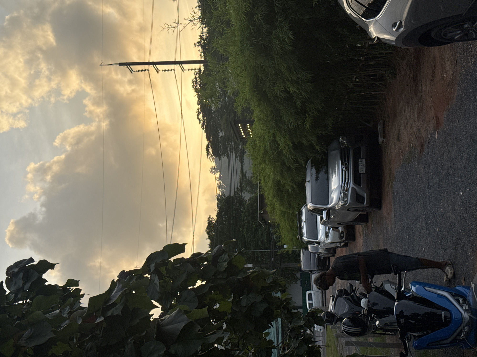 The image shows a street scene in Wichit, Thailand, during what appears to be sunset or dusk. A person is standing next to several motorcycles, some of which are parked on a gravel surface. Behind the motorcycles, a line of cars is parked along the side of the road, including a silver SUV and a white pickup truck. A dense hedge of bamboo lines the right side of the road, and lush green trees are visible in the background, with a hillside beyond. Overhead, telephone poles and wires are silhouetted against a sky filled with warm-toned clouds, suggesting either sunrise or sunset. The overall lighting is dim, consistent with the end of the day.