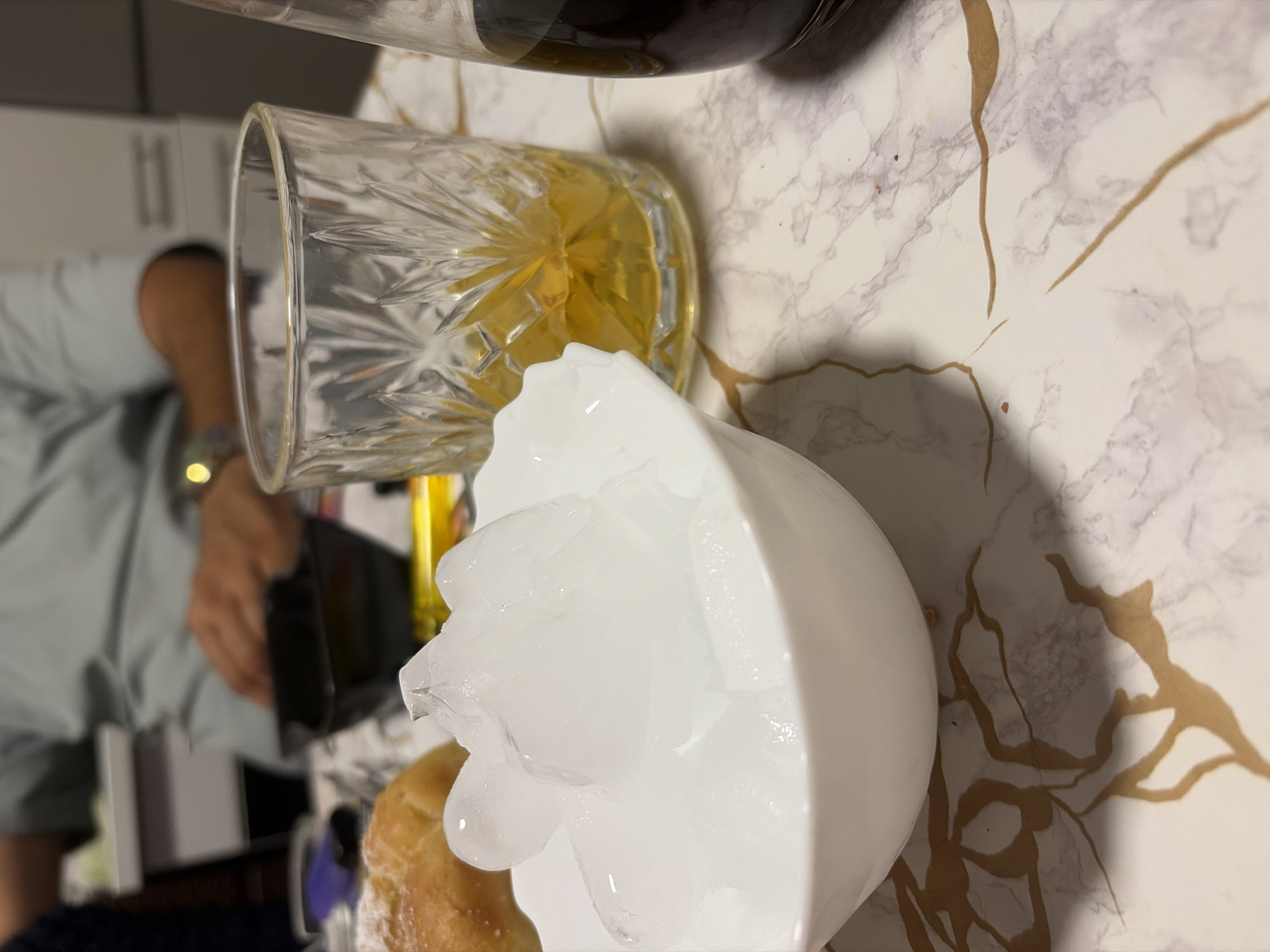 In a dining setting, possibly a home in Duisburg, Germany, a person in a light blue shirt is seen with their hands in a black bowl. In the foreground, a white bowl is filled with ice cubes. To the right of the ice bowl is a clear glass with an amber liquid, possibly juice or a spirit. Beside that, a dark liquid is visible in a glass bottle. The table has a marble-like surface with gold veins. A baked good, possibly a pastry, is partially visible to the left of the ice bowl. The overall lighting suggests an indoor environment, possibly a meal or a gathering.