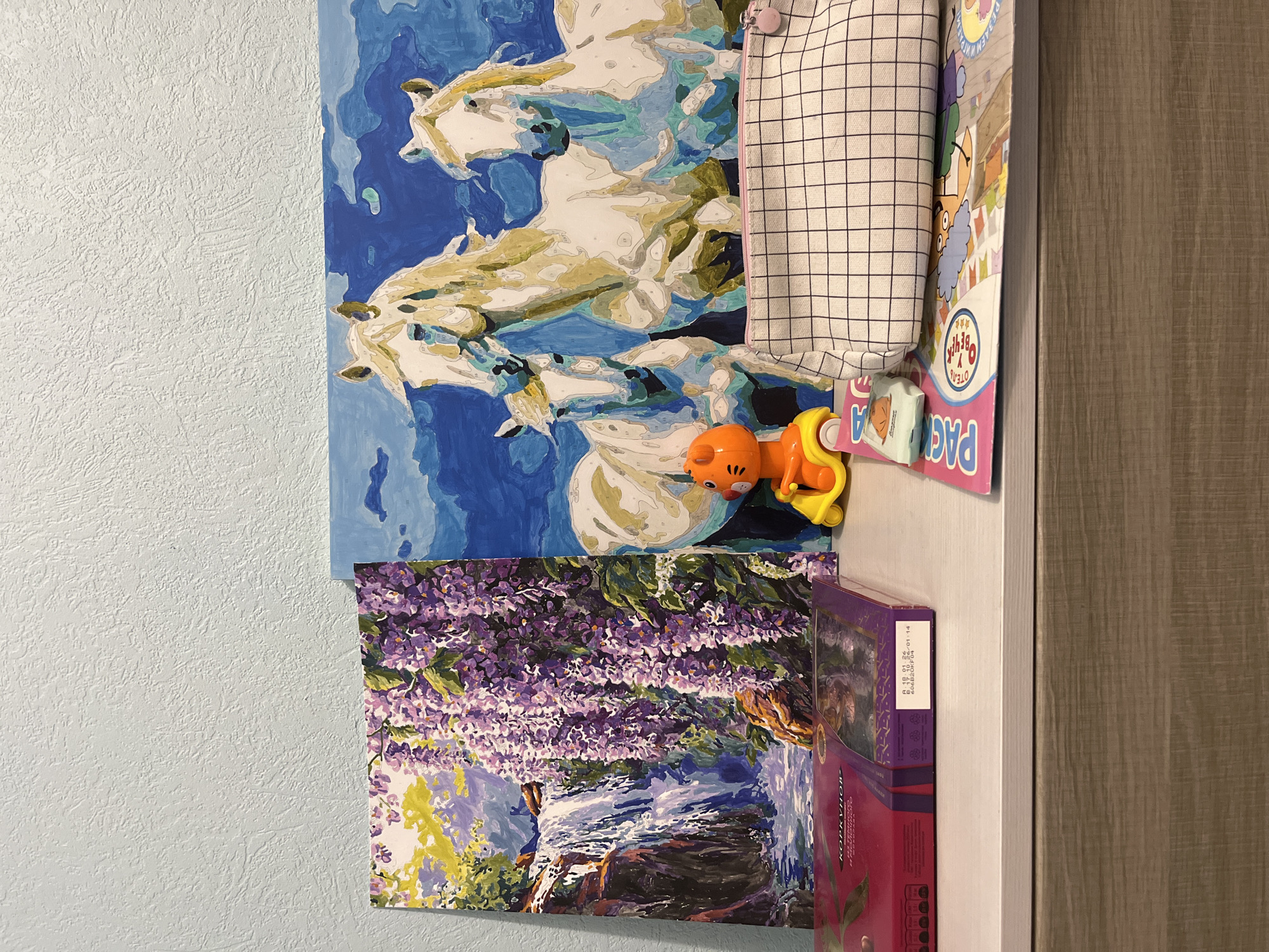 The image shows a still life of objects against a pale blue wall with a textured surface. In the upper part of the image, there's a painting depicting three white horses set against a blue background. Adjacent to it, on the right, is a white zippered pencil case with a black grid pattern and pink trim. Below it sits an orange and yellow toy. Underneath the horse painting is another painting, this one featuring purple wisteria cascading over a scene with a waterfall or flowing water. To the right of the wisteria painting, there's a box of chocolates with visible Russian text "КОРКУНОВ" and expiration date information, which suggests the location could be in Russia.
Based on the brand of the chocolate box "Korkunov" and the language used on it, the location is likely in Russia. Additionally, the toy packaging has text in Russian. Without further information, it's not possible to pinpoint the exact location as Izhevsk.