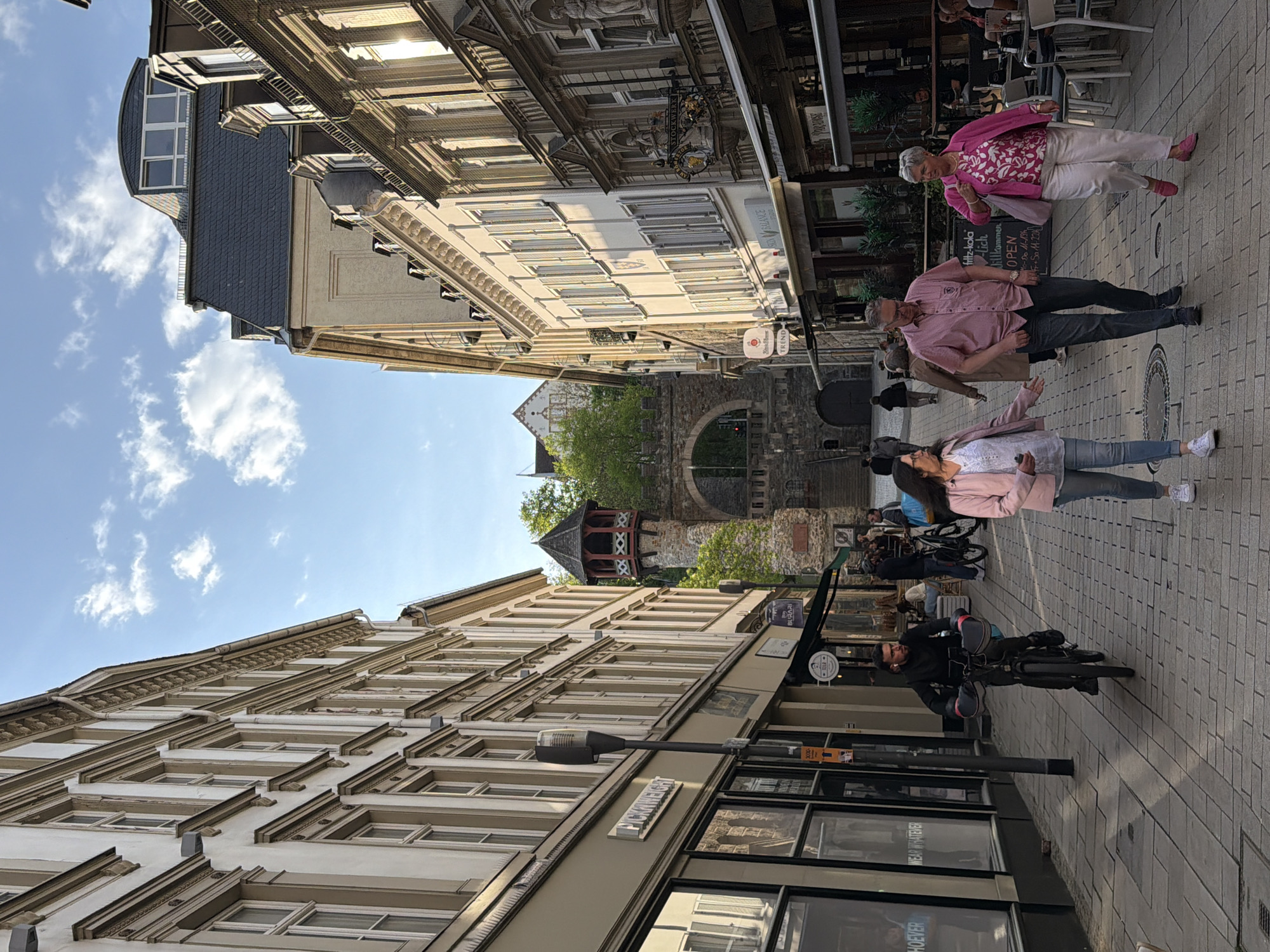 The image depicts a street scene in Wiesbaden, Germany, showcasing a narrow pedestrian area between tall buildings. The sky is visible above, partly obscured by the buildings.
Several individuals are present: An older woman wearing a pink jacket and white pants is walking toward the top-right corner. A person in a pink shirt and dark pants is walking close to her. Another woman wearing a pink jacket and jeans is walking and appears to be gesturing with her arm. A person on a bicycle is visible along the lower section of the image.
Architectural details include a tower visible at the end of the street. Various shops and stores with signs are also visible along the street. A sign on the left reads “HOEVER”.
The street is paved with rectangular blocks. There is outdoor seating associated with a business.