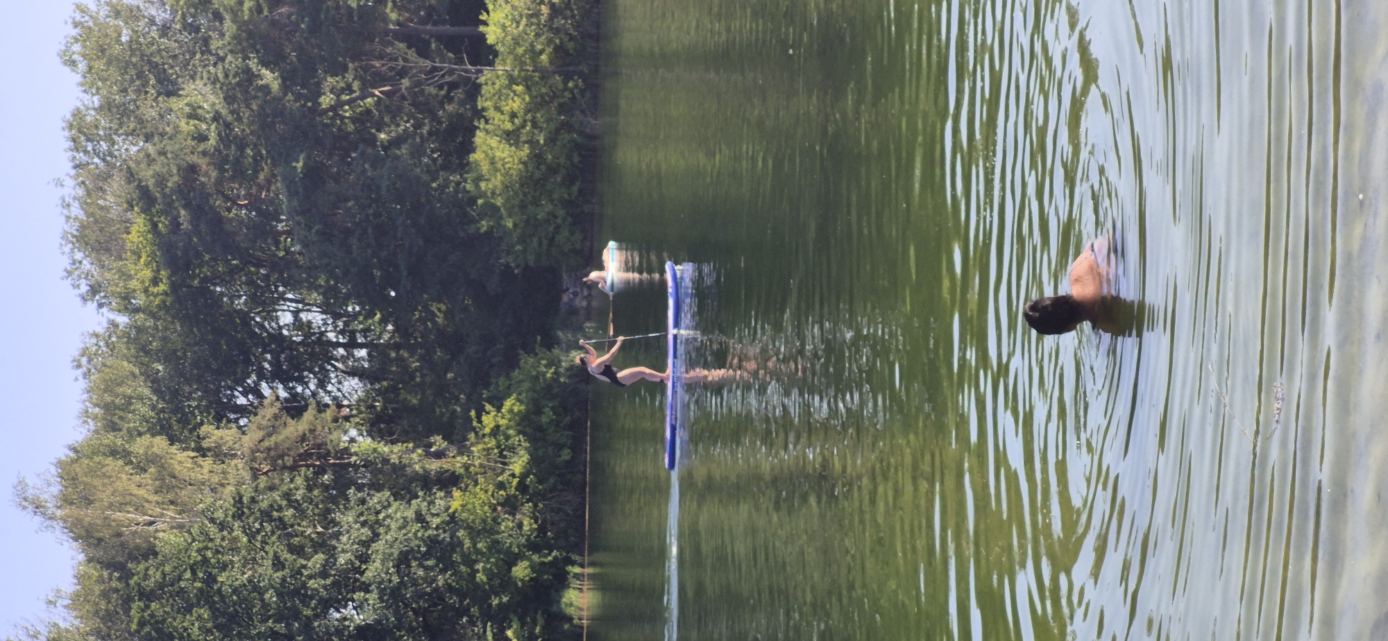 The image depicts a serene lake scene surrounded by lush green trees. In the foreground, a person is swimming, with their head and shoulders visible above the water, creating concentric ripples. Further in the background, another individual is paddleboarding on a blue inflatable board, holding a paddle. Behind them, a third person is partially visible, possibly sitting or lying on a light-colored inflatable. The water is calm and reflects the surrounding foliage and the blue sky. The trees form a dense backdrop, suggesting a natural setting. There is no visible indication of this location being in Ingolstadt, Germany.