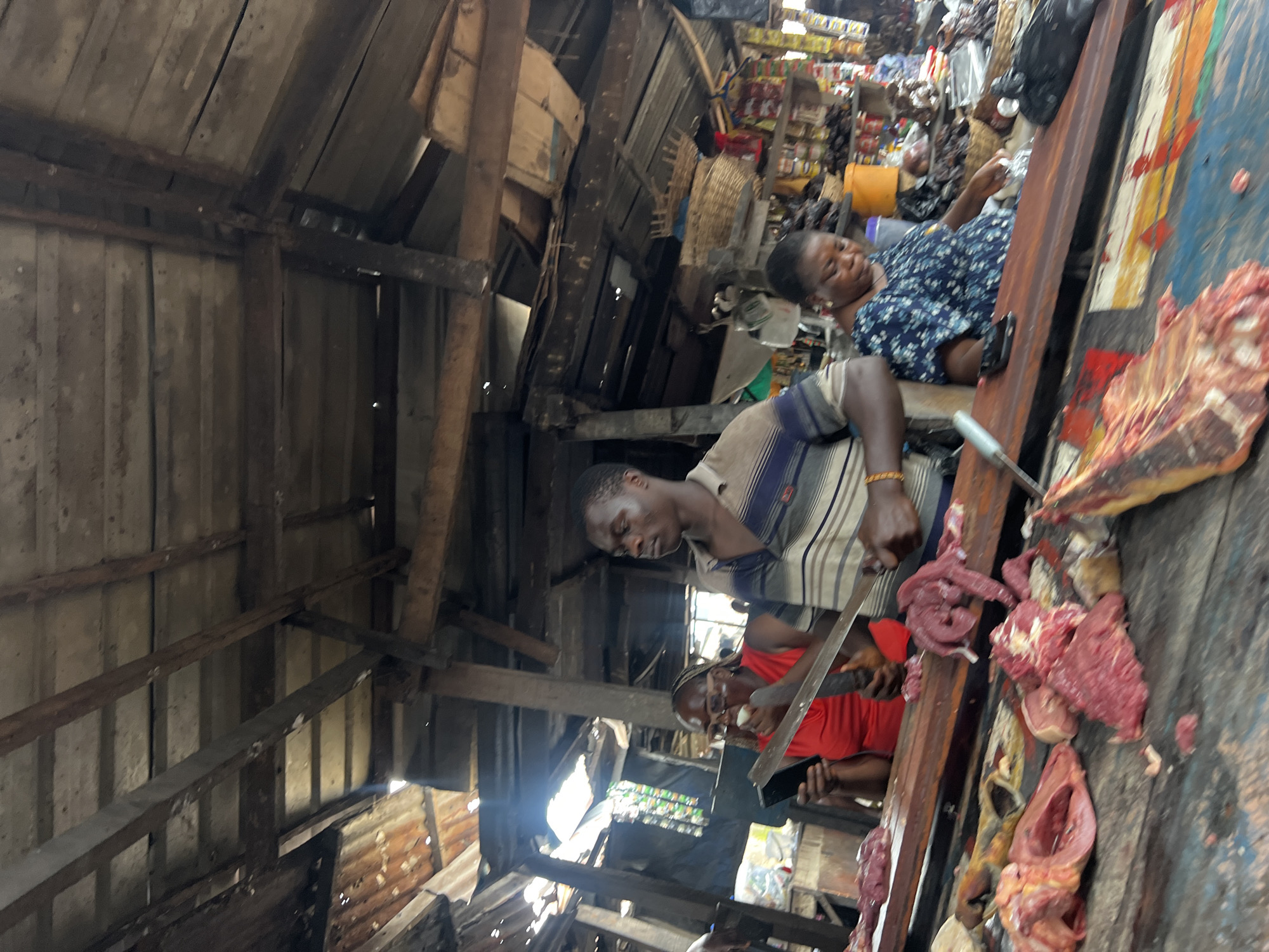 The image depicts a busy market scene in Nkpor, Nigeria. In the foreground, a man wearing a striped shirt is seen preparing meat. He is holding a large knife and appears to be actively cutting or dividing a piece of raw meat. Several other people are visible in the background, including a woman in a blue patterned dress lying down, possibly resting, and another person in a red shirt. The setting is an open-air market with a rustic, wooden structure providing shelter. The market appears crowded and filled with various goods, suggesting a typical day of commerce. Raw meat is prominently displayed on a wooden surface, indicating a butcher's stall or a section of the market dedicated to selling meat. The overall atmosphere seems lively, with multiple individuals engaged in their respective activities. The lighting suggests it is daytime.
