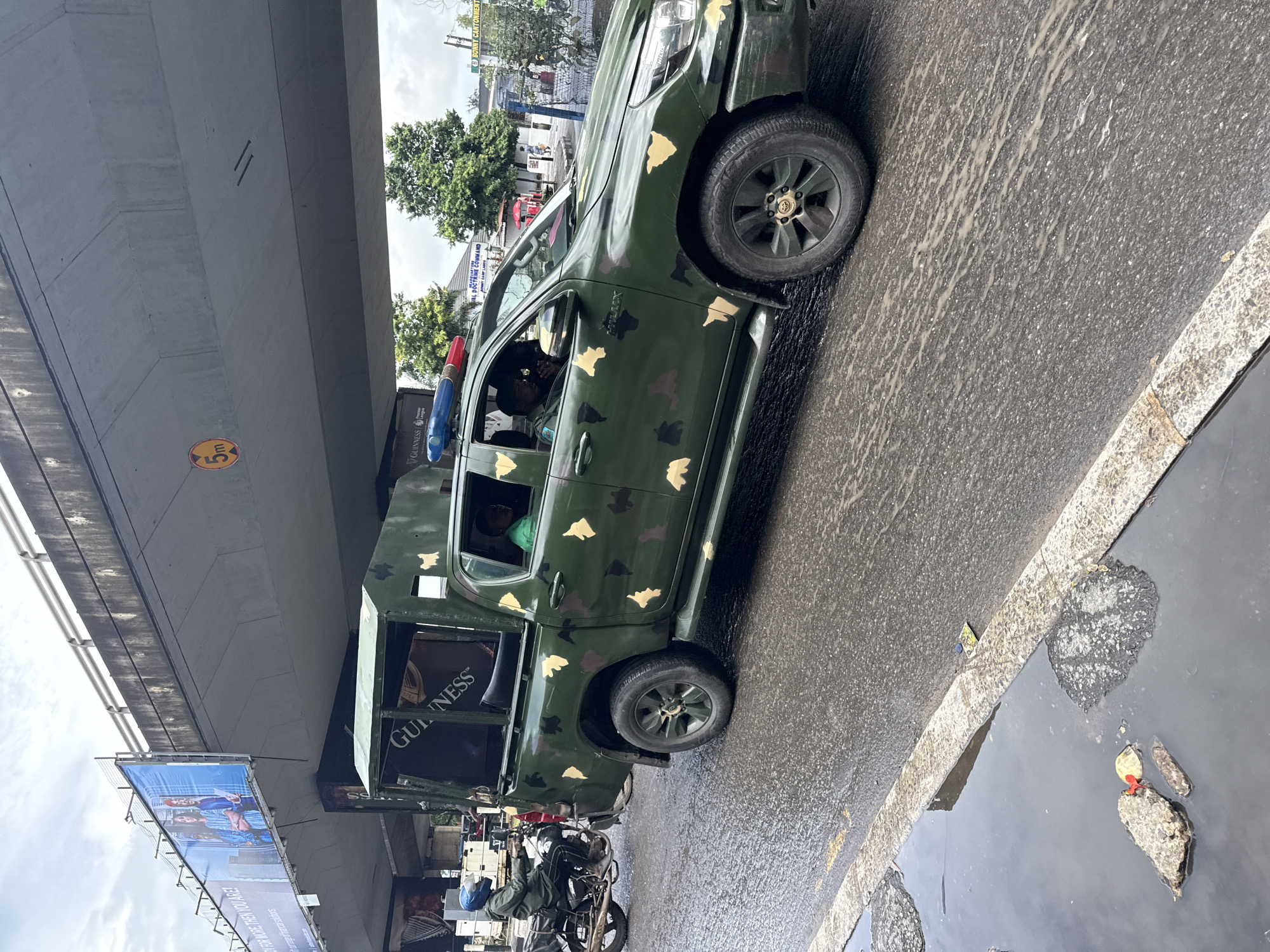 This image depicts a scene in Ikoyi, Nigeria, likely during the day under overcast skies, as indicated by the wet road and reflections. The primary focus is a green, camouflage-patterned pickup truck, likely a security vehicle, with a red and blue light bar mounted on its roof. The truck has a reinforced rear section, possibly for carrying personnel or equipment. The side of the truck features a prominent "GUINNESS" logo, suggesting a sponsorship or advertising presence.
In the foreground, a motorcycle with a rider wearing a blue helmet is partially visible, hinting at the common mode of transport in the area. The road surface appears wet and somewhat dirty, with visible tire tracks and scattered debris. Overhead, a large concrete overpass or bridge dominates the upper portion of the frame, casting a shadow. Below the overpass, a billboard advertising "FOR MORE THEN YOU ARE!" with images of people is mounted. Another sign for "BONNY CANTONMENT" and "NAVAL DOCTRINE COMMAND" is visible on a building in the background, suggesting a military or naval presence in the vicinity. The overall atmosphere appears to be a busy urban environment, possibly during or after a rainfall.
