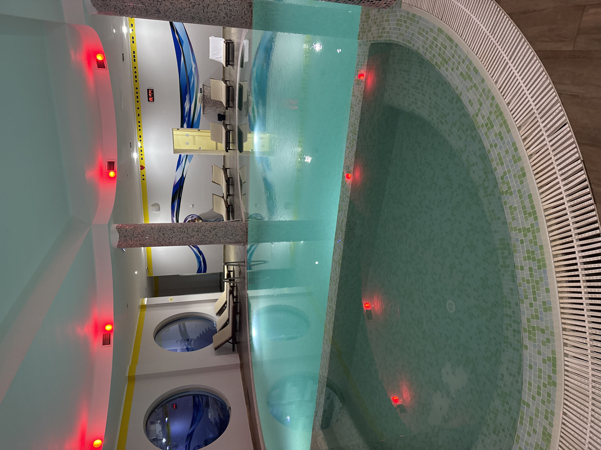 The image depicts an indoor swimming pool complex, featuring a long rectangular pool connected to a circular hot tub. The rectangular pool is filled with turquoise water. The hot tub is tiled with small green and blue tiles. Along the wall are reclining chairs and round windows. Red lights are visible along the pool and walls. The specific city and location within Unknown, Unknown, are not discernible from the image. There are no people visible in the scene.