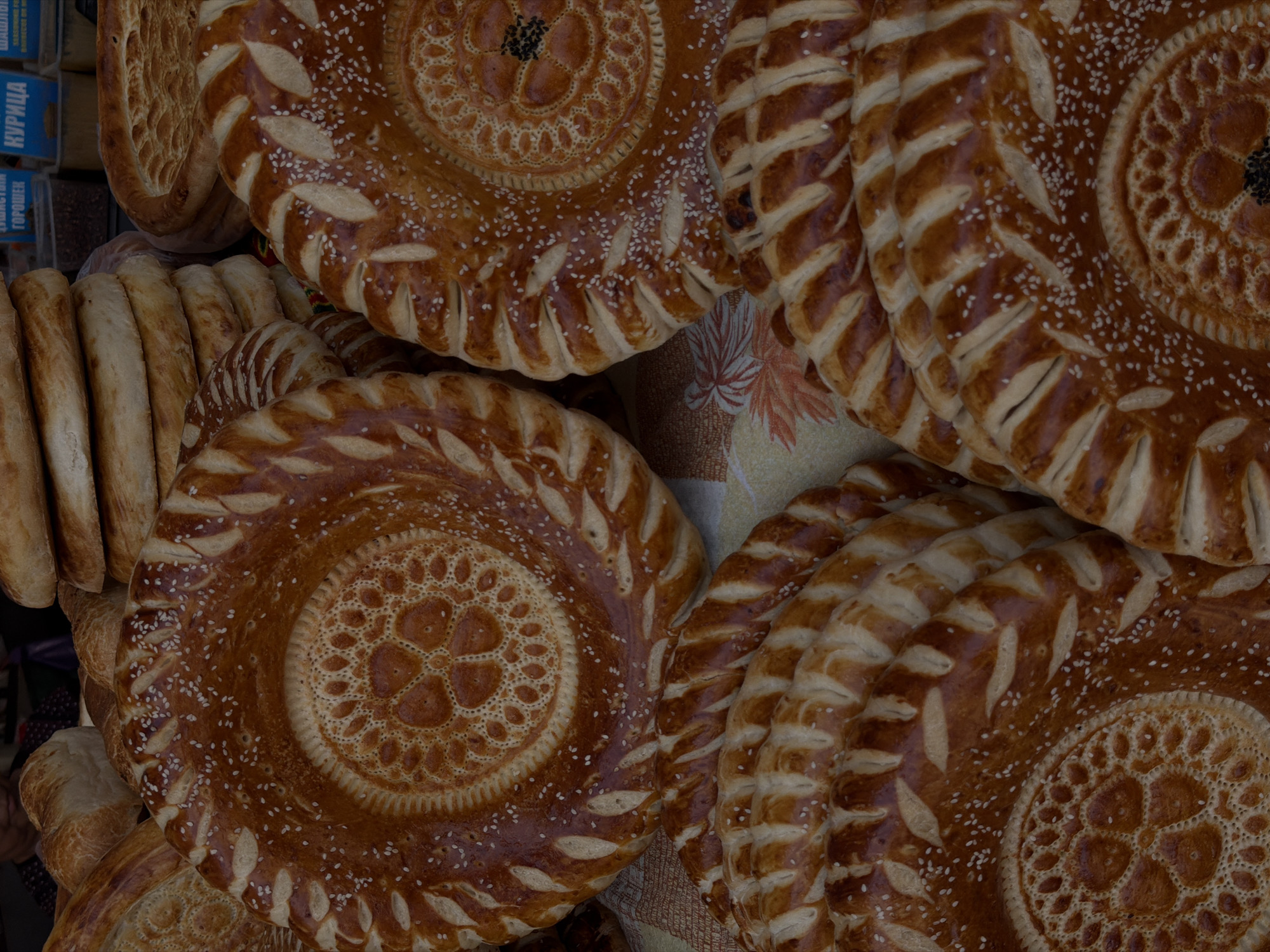 A close-up, vertical, eye-level shot displays a variety of round, decorative flatbreads piled on a table. The breads are a golden-brown color, with some appearing lighter and others darker, indicating variations in baking. Each bread features a prominent, intricate circular pattern in the center, surrounded by a decorative border of carved leaf-like shapes. Many of the breads are sprinkled with white sesame seeds. In the background, out-of-focus loaves of bread are stacked, and partially visible signage in a foreign language can be observed, suggesting a market or bakery setting. A portion of a person's shoulder and head is visible in the upper left corner. The table is covered with a patterned cloth that has floral motifs in shades of yellow and orange. The lighting is soft and natural, casting subtle shadows.