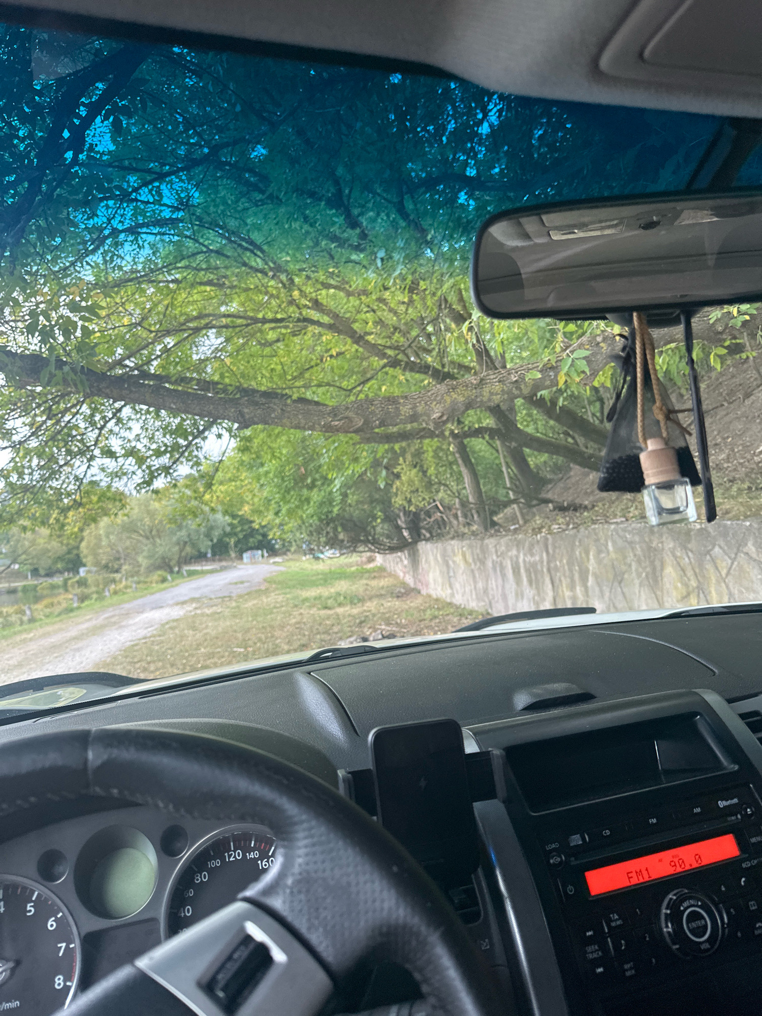 The image is a first-person view from the driver's seat of a car. The car's dashboard and steering wheel are in the foreground. The dashboard features a radio display showing "FM1 90.0", a phone mounted on a stand, and part of the instrument cluster including a speedometer that reads approximately 50 km/h. Outside the car, visible through the windshield and side windows, is a tree-lined dirt road leading away from the viewer. The branches of a large tree hang low over the road and the car. The environment appears to be rural or park-like, with green foliage and a glimpse of water to the left. The scene is set in Makiv, Ukraine.