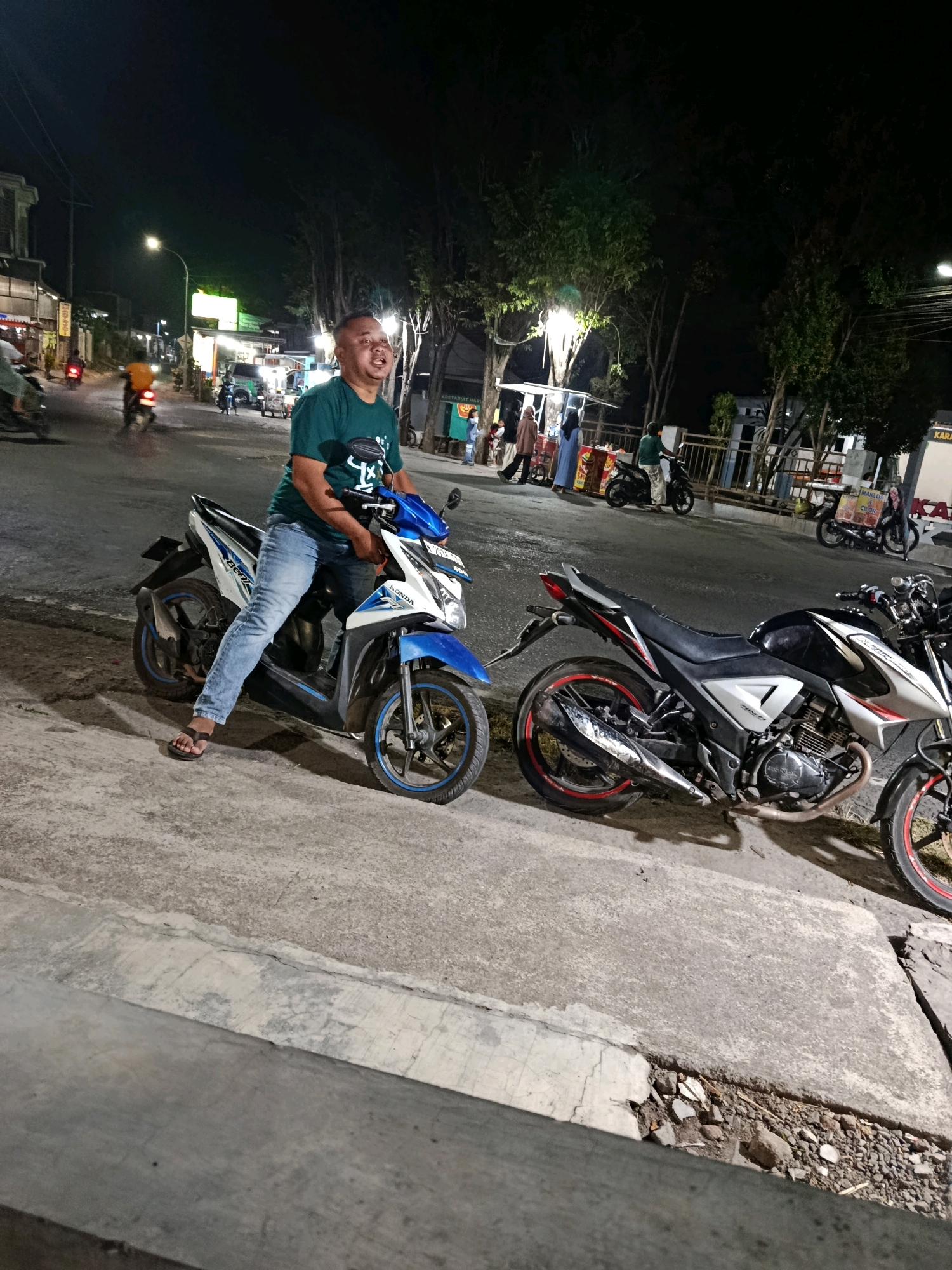 A man in a green t-shirt and blue jeans is sitting on a blue and white Honda Beat motorcycle at night. The motorcycle is parked on the side of a street, likely in Pager, Indonesia. The man is looking to his left and appears to be talking. In the background, there are other motorcycles, streetlights, trees, and a few people walking. The scene appears to be a busy street with food stalls or shops lining one side. Another motorcycle, a black and white Honda CB, is parked next to the man's motorcycle.