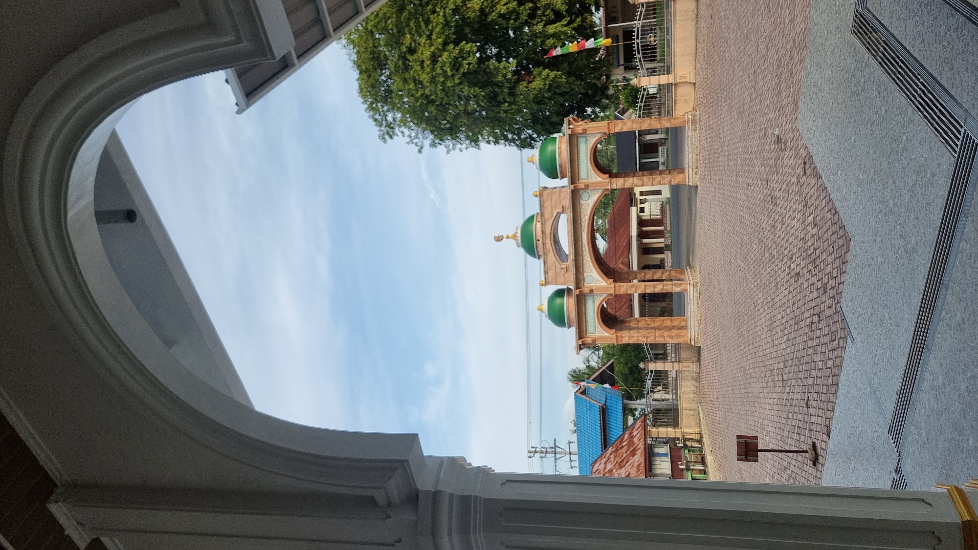 The image shows an arched entrance with three green domes and a crescent moon at the top, likely a mosque or religious building. The entrance is made of stone or brick. It is situated in an open paved area. In the background, there are several buildings with red and blue roofs, and a large tree with lush green foliage. A flag with red, white, and green stripes is visible near the entrance. The sky is partly cloudy and light blue. The foreground features a white arched structure framing the view, suggesting the image is taken from inside a building. A small wooden signpost stands on the paved ground to the left. The scene appears to be a public space in Meunasahlhok, Indonesia, possibly during daylight hours, given the lighting.