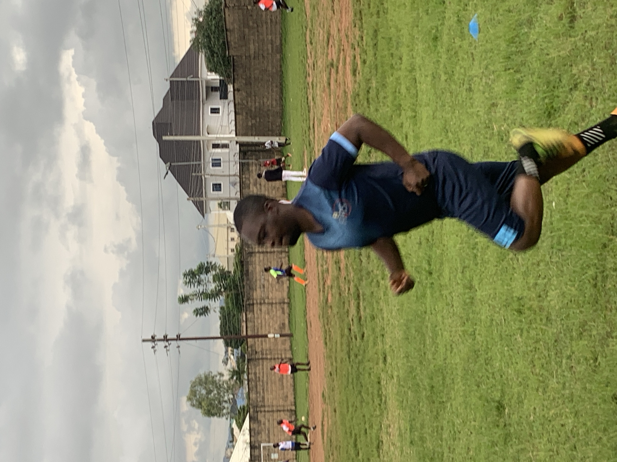The image depicts a soccer training session on a grassy field in Jos, Nigeria. The foreground features a Black man in a blue soccer uniform with light blue accents, actively running. He is wearing dark blue shorts with a light blue stripe and black and white striped socks with yellow soccer cleats. In the background, other individuals in soccer attire are visible on the field. A brick wall runs along the perimeter of the field, and beyond that, modern buildings, including a large house with a brown, angular roof, and utility poles with wires are present. The sky is overcast with heavy, gray clouds, suggesting a cloudy day. The overall setting appears to be an outdoor sports facility, possibly within a residential or semi-urban area of Jos.