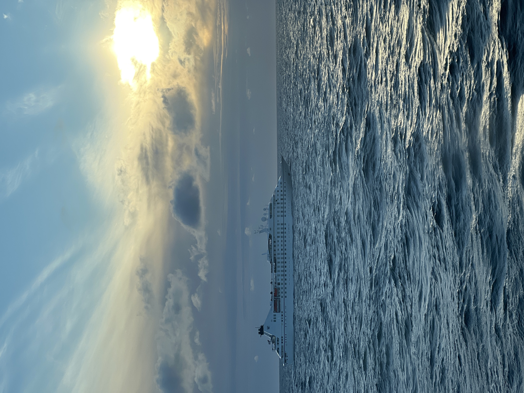 An exterior, eye-level, medium shot captures a white cruise ship sailing on a choppy, dark blue sea under a partly cloudy sky. The sun is a bright, yellow orb in the upper right corner, partially obscured by clouds and casting a golden glow on the scene. The ship, positioned in the middle left of the frame, has multiple decks and appears to be a passenger vessel. The water is textured with small waves, reflecting the light from the sky. The sky transitions from a pale blue at the top to a darker, more muted tone near the horizon, with streaks of white and grey clouds. The location is not specified in the image.