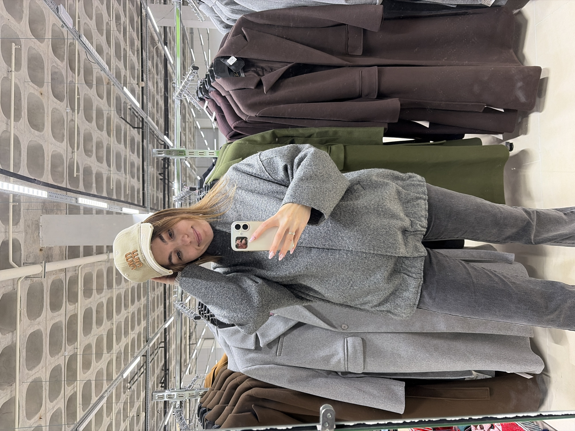 A young woman is taking a selfie in what appears to be a clothing store in Izhevsk, Russia. She is wearing a grey sweater, grey jeans, and a beige baseball cap. She is holding a smartphone in her right hand. Behind her, there are racks of coats in various colors including brown, green, and grey. The wall in the background is made of concrete blocks with circular openings. The scene is well-lit by fluorescent lights.