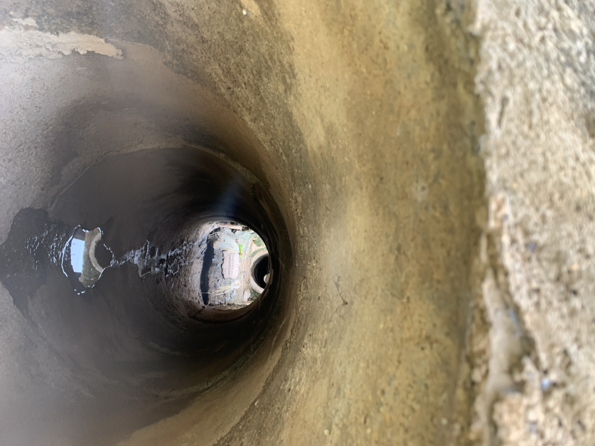 The image presents a longitudinal view from within a large, circular concrete drainage pipe, looking towards its exit in Rafin Par, Nigeria. The interior surface of the pipe is rugged and textured, displaying a gradient of earthy tones from light tan to dark grey, consistent with accumulated sediment and moisture. A narrow stream of water flows along the invert (bottom) of the pipe, collecting in a small, reflective puddle towards the foreground, which mirrors a brighter area at the pipe's terminus.
At the far end of the primary pipe, a smaller, dark, circular opening, possibly another conduit or a break in the pipe, frames a view of the external environment. Beyond this opening, an uneven ground surface composed of soil and debris is visible, along with sparse green vegetation. Positioned on the distant ground is a light-colored, rectangular slab or plaque, bearing the inscribed word "WELCOME." No individuals, animals, or specific interactions are depicted within the scene, which portrays a segment of drainage infrastructure in an outdoor setting.