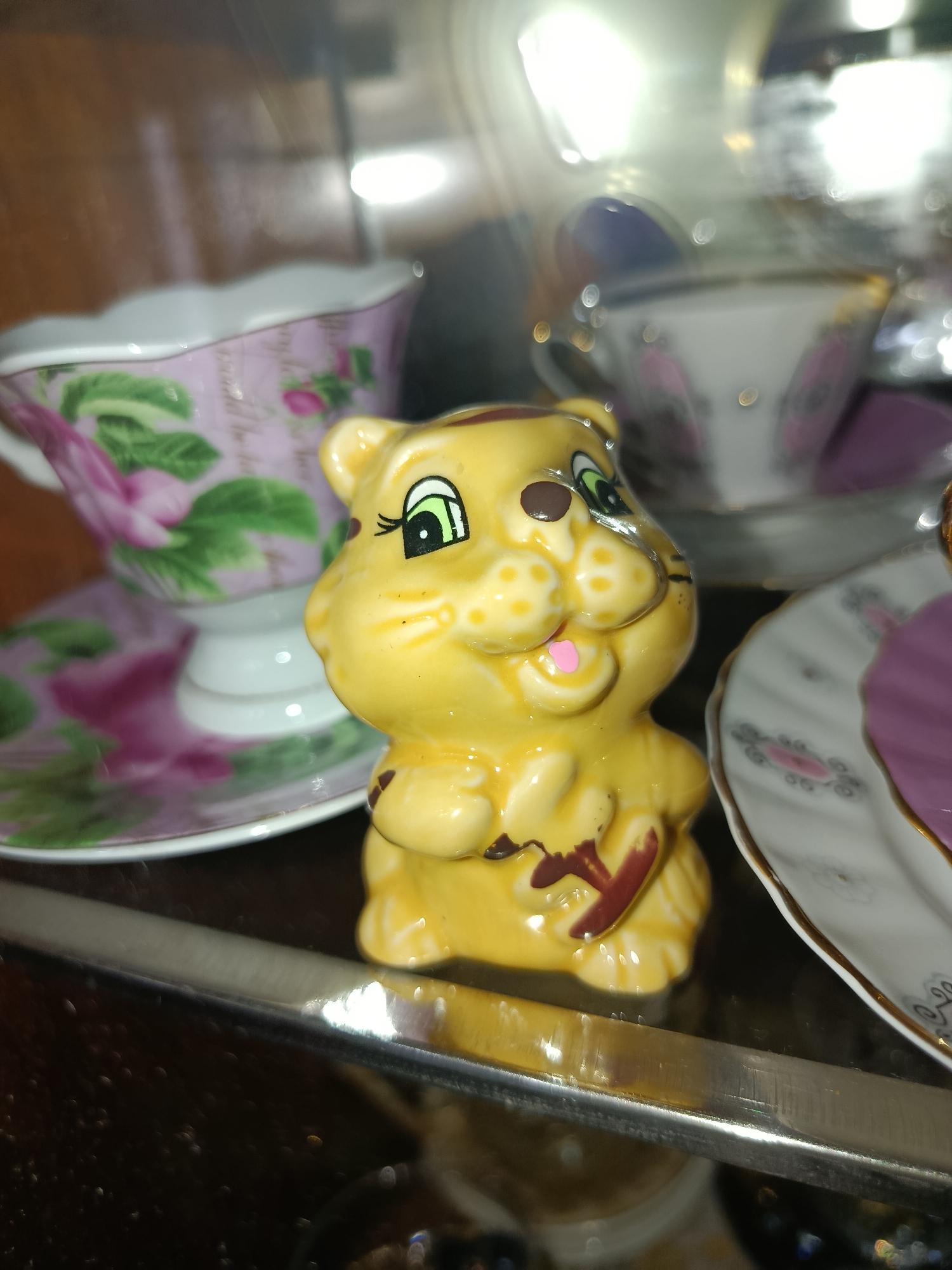 A yellow ceramic figurine of a cartoonish cat, with large green eyes and spots on its face, sits in the foreground. The cat figurine is holding something brown in its paws. Behind the cat, a delicate porcelain teacup and saucer with a floral design are visible. Further in the background, other pieces of ornate porcelain tableware are partially obscured. The scene appears to be an indoor display, possibly in a cabinet or on a shelf, with reflections from lighting and glass surfaces. The location is not determinable from the image provided.