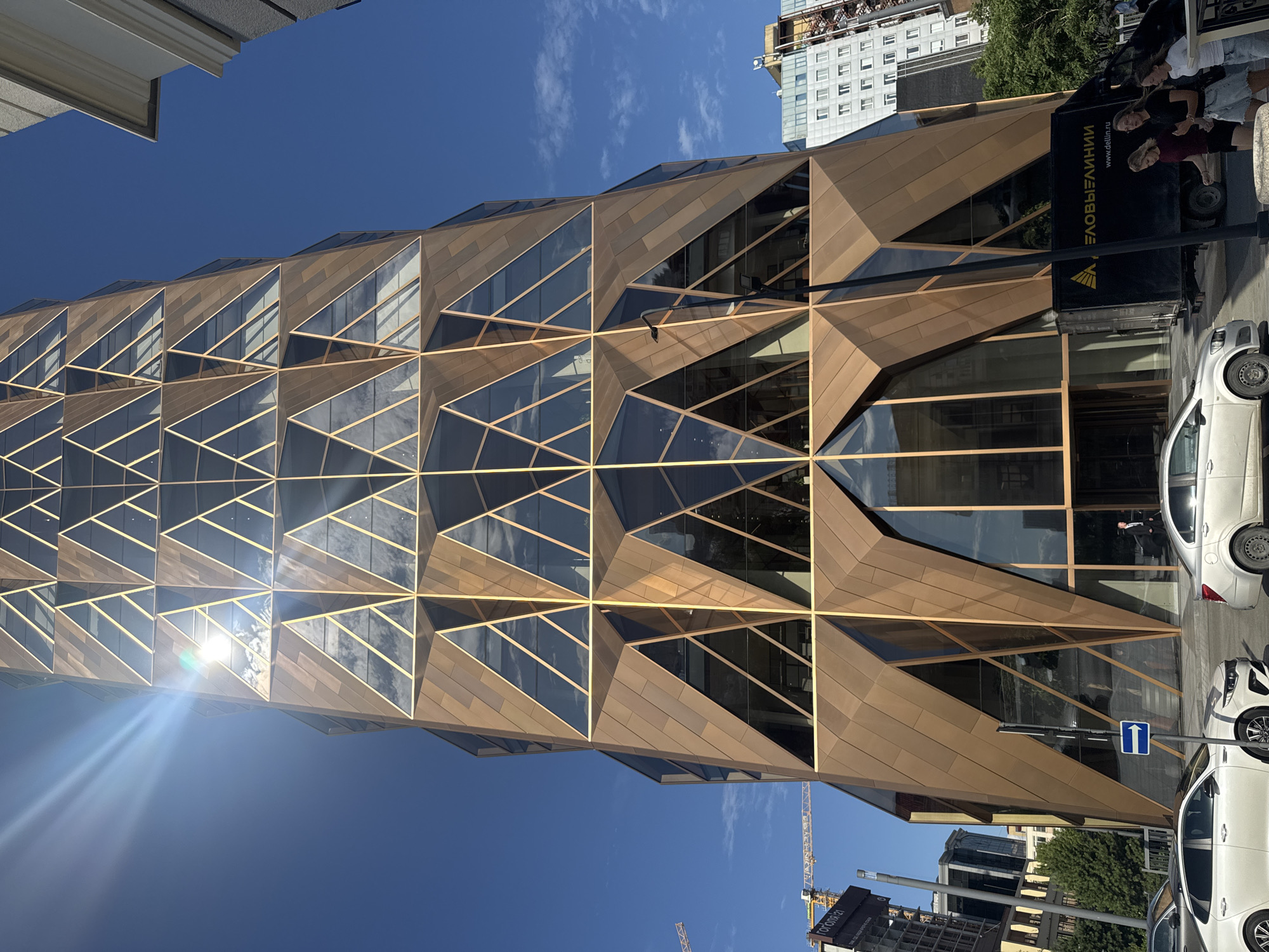 The image shows the skyscraper Vysotsky in Yekaterinburg, Russia, on a clear, sunny day. The skyscraper's facade is a distinctive geometric pattern of bronze-colored panels and glass windows. Sunlight reflects strongly off the windows and the building's surface, creating lens flare and bright highlights. In the foreground, a street is visible with several cars, including a white sedan on the left and a silver sedan on the right. A black delivery truck with the text "ДЕЛОВЫЕ ЛИНИИ" (Delovye Linii) on its side is parked near the building's entrance. Three young women are walking on the sidewalk in front of the truck. A street lamp and a directional sign pointing right are also present on the sidewalk. In the background, other buildings and construction cranes can be seen, indicating an urban environment. The sky is a vibrant blue with a few wispy clouds.