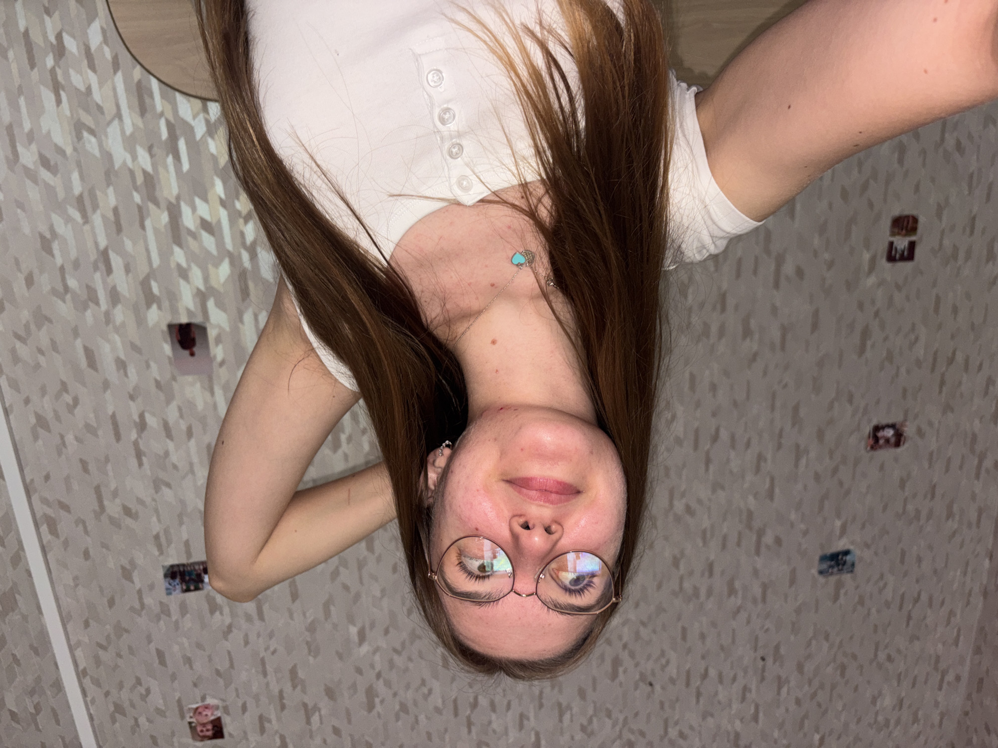 An indoor portrait photograph captures a young woman from an inverted perspective. She has long, straight, reddish-brown hair that frames her face and shoulders, which are clad in a white short-sleeved top with a button placket. Around her neck is a delicate silver chain with a small, light blue heart or turtle-shaped pendant and another tiny silver charm. She wears round, thin-rimmed glasses, through which her eyes reflect ambient light. Her expression is calm, with a subtle, gentle smile. Her skin shows natural texture and minor blemishes. One arm is visible, bent behind her head.
The background is a wall adorned with a repeating geometric pattern of light brown, beige, and white diamond or arrow shapes. Several small, rectangular photographs are casually taped to the wall, hinting at a personal space like a bedroom. The lighting is soft and even, typical of indoor artificial light. The image lacks any discernible text. This scene is set in Perm, Russia.