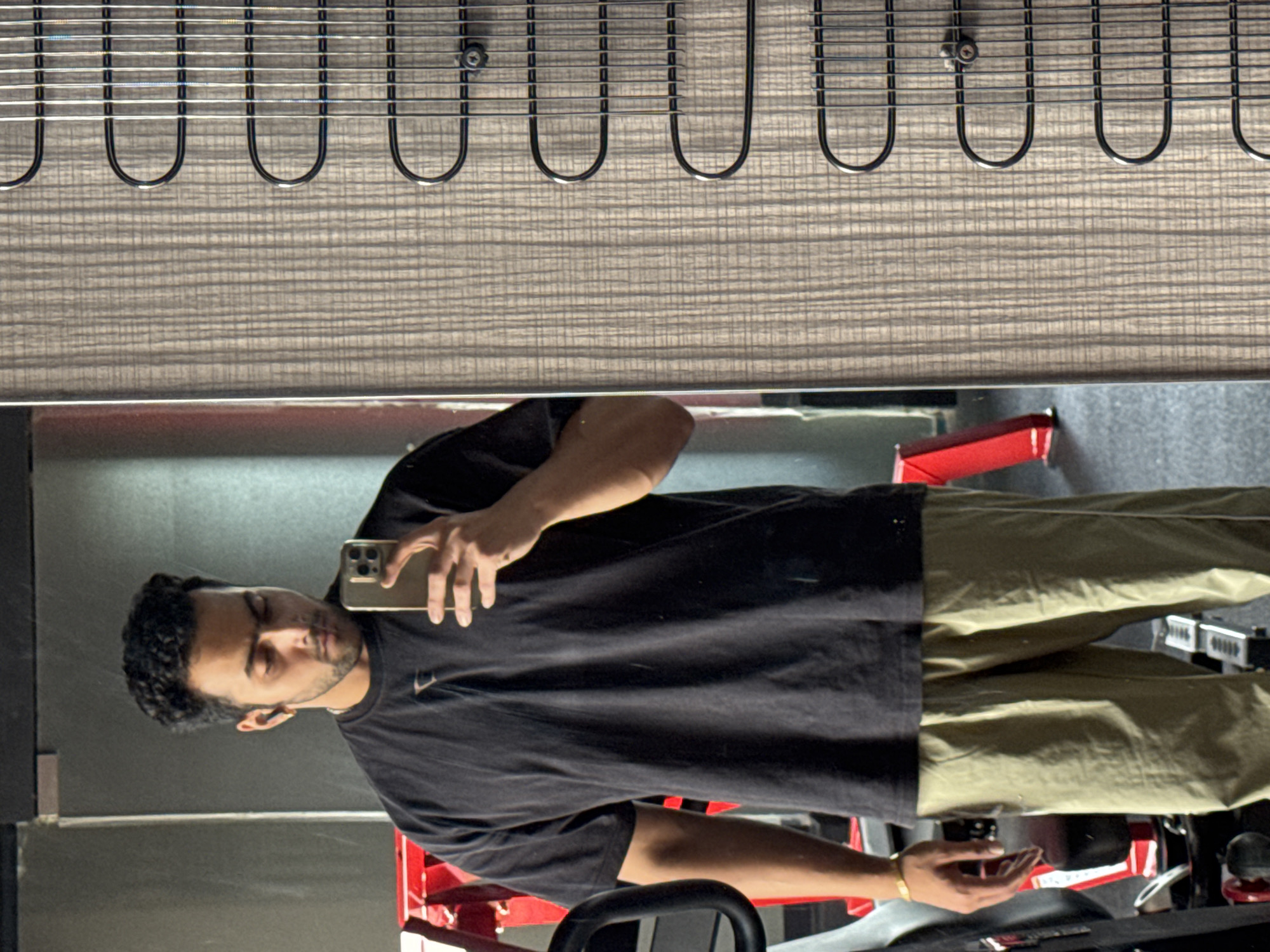 The image shows a young man taking a mirror selfie in a gym. He is wearing a black t-shirt and khaki pants. He has short, curly dark hair and is wearing wireless earbuds and a gold bracelet. The gym appears to be equipped with weight machines, as indicated by the red equipment visible in the background. The man is holding a smartphone in his right hand, presumably to take the photo. The background also features a textured wall with a pattern of vertical lines and horizontal curves, possibly a decorative element or part of a locker or appliance. The overall lighting suggests an indoor environment, consistent with a gym setting. The location is not specified in the image and cannot be determined.