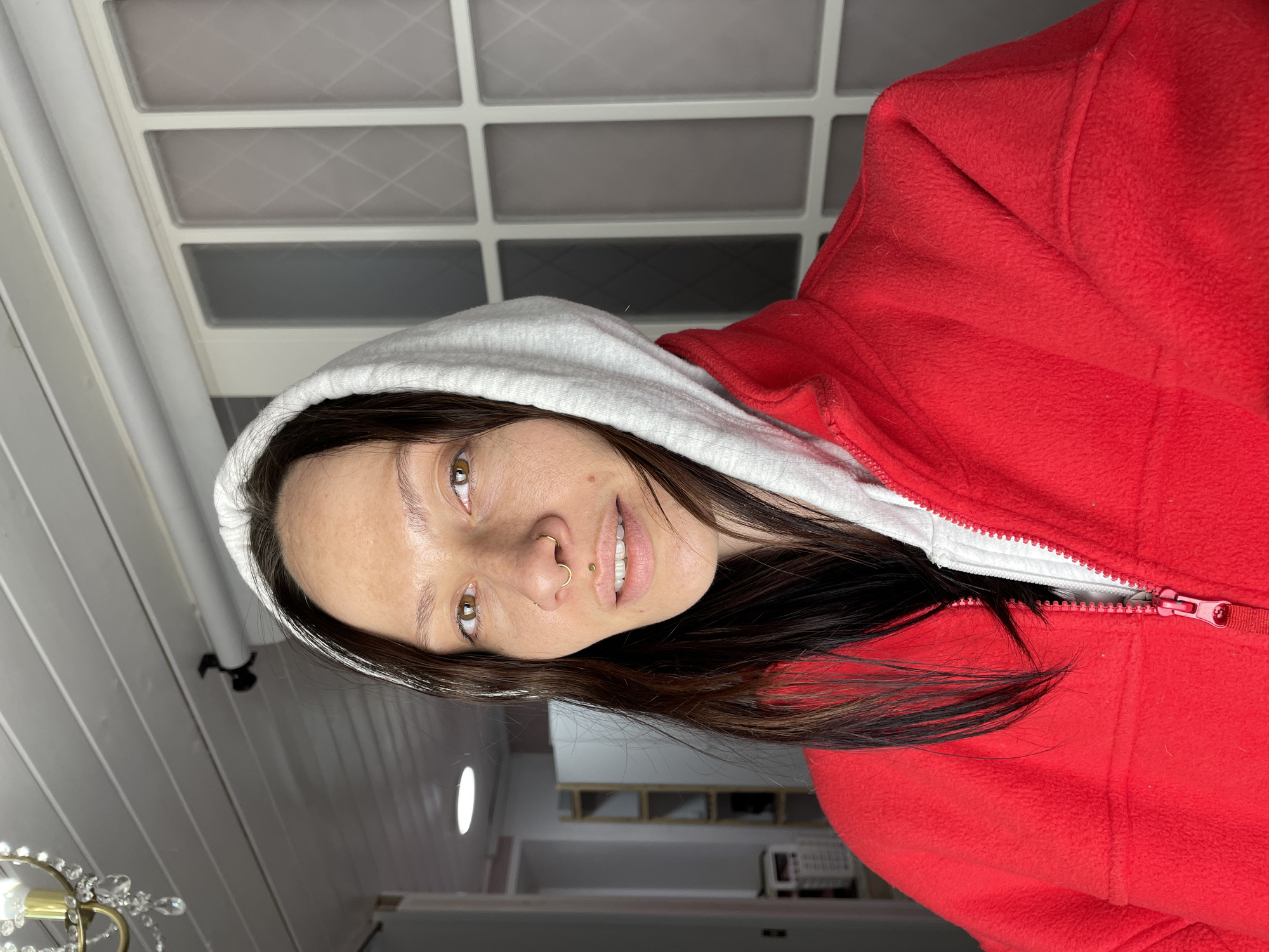 A young woman is photographed indoors. She wears a red fleece jacket with a white hood. Her dark hair falls around her shoulders. She has light skin, brown eyes, and piercings in her nose and lip. The background is blurry but shows a light-colored room with shelves. It is not possible to identify the location as Izhevsk, Russia, from the given visual information.