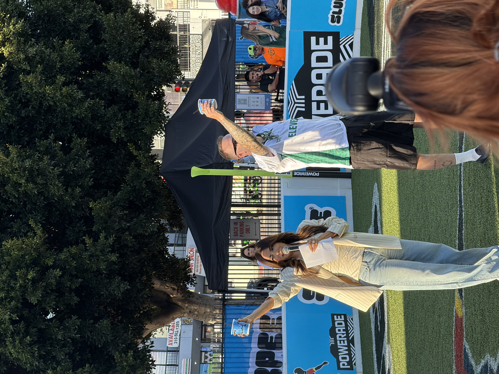 The image depicts an outdoor event, likely a promotional gathering, set on a vibrant green turf field. In the foreground, a woman with long, dark, wavy hair is speaking into a microphone, holding a white paper in her left hand and a blue cup in her raised right hand. She is wearing a striped blazer over a light-colored top and light-wash jeans. To her right, a man with short hair, sunglasses, and tattoos on his arm is also holding a blue cup aloft in his right hand. He is wearing a white t-shirt with green accents and black shorts.
Behind them, a black tent is set up, and further back, there are buildings with visible signage. One sign reads "AVAILABLE" with a phone number, another "HAIR SALON," and a third appears to be "BRIENTOS." In the background, several people are visible, some standing behind a blue barrier, others further back near the buildings. A traffic light with red illumination is also present in the background. The overall atmosphere suggests a sunny day in an urban environment. The presence of "POWERADE" branding on banners and flags indicates a sponsorship or collaboration with the sports drink company. The style of the buildings and the general ambiance suggest a location within the city of Los Angeles, United States of America.